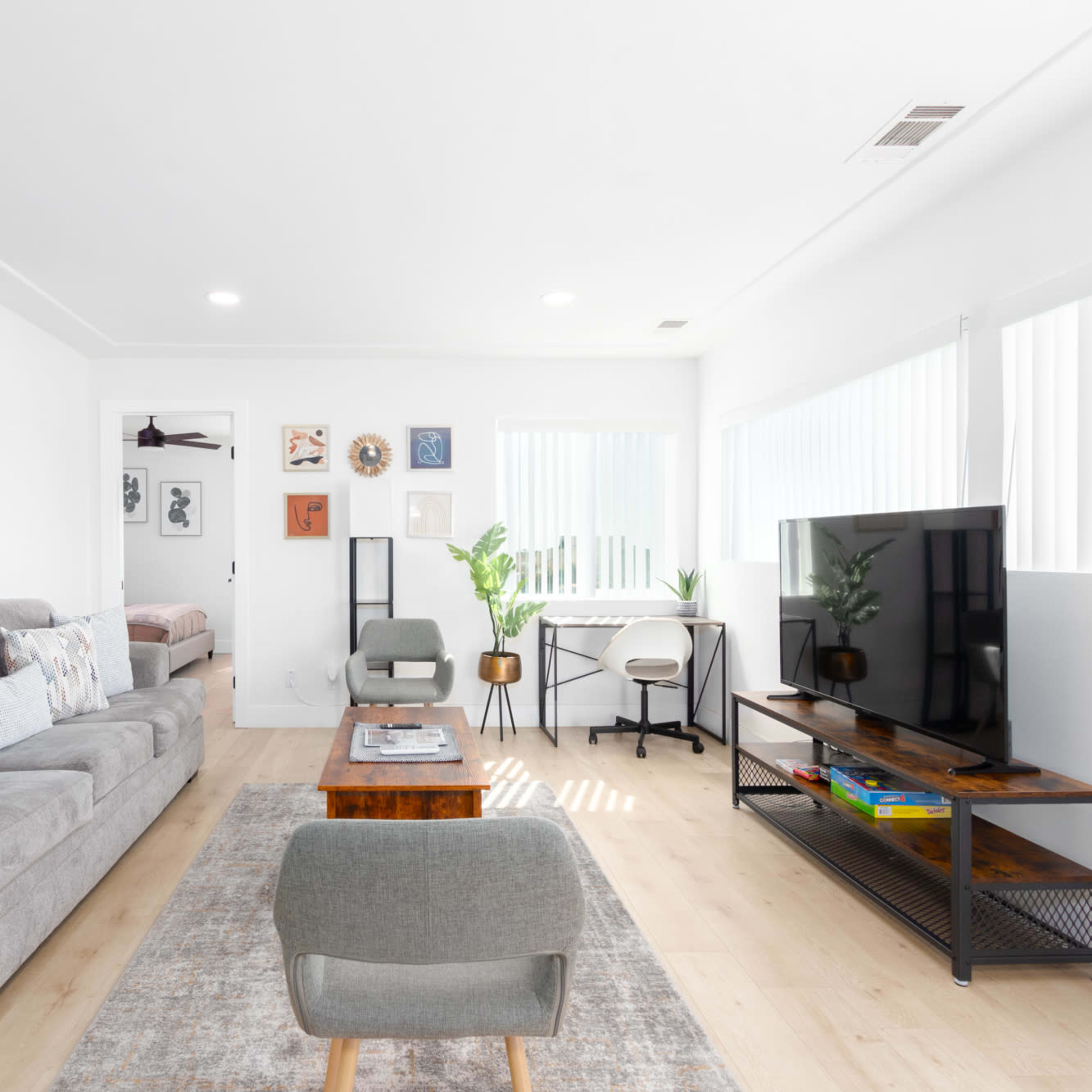A modern living room features a gray sectional sofa, a wooden coffee table, a mounted TV, and large windows with blinds, framed by abstract wall art.