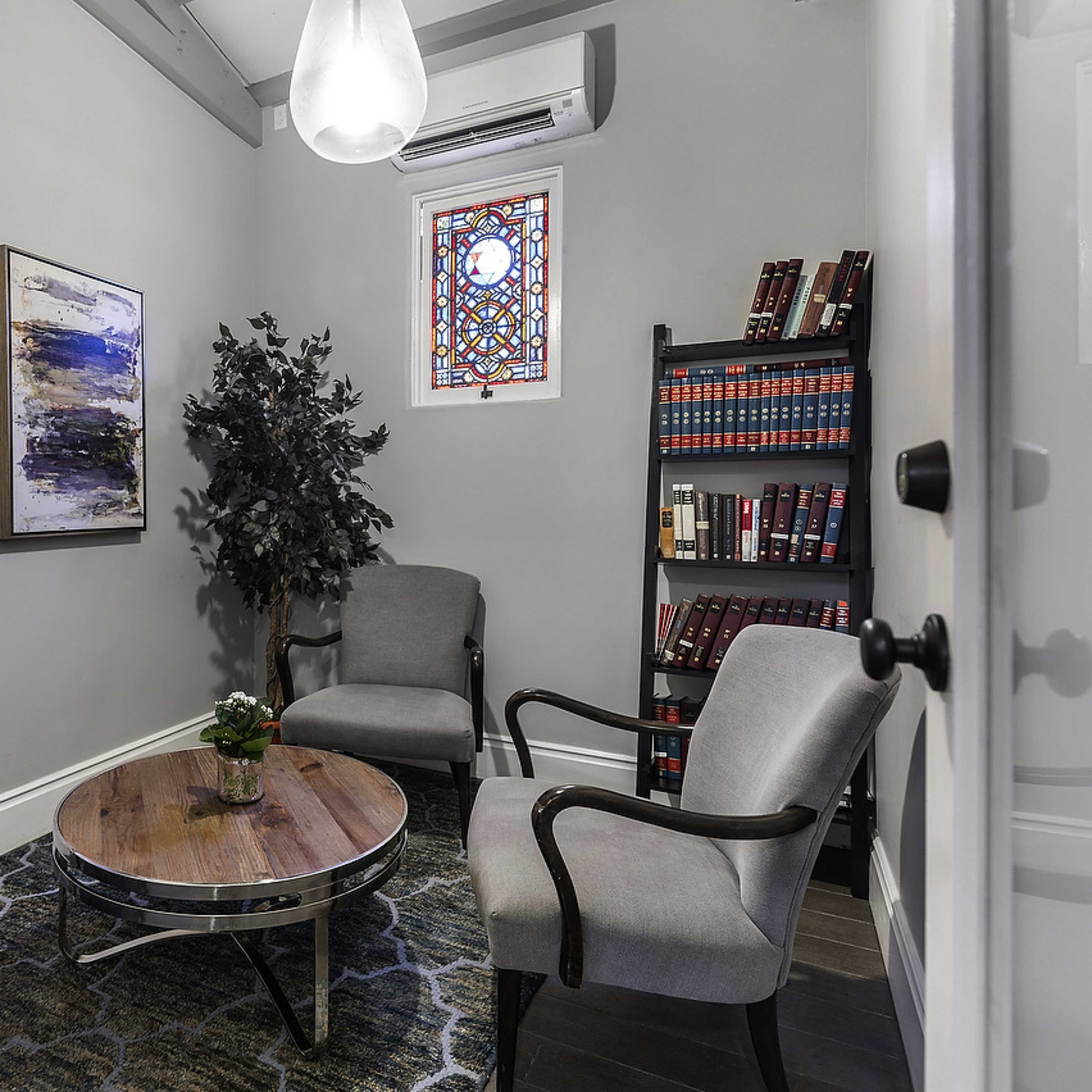 A small, well-furnished room features two chairs, a round wooden table, a potted plant, a bookshelf with books, and two framed artworks on the walls.