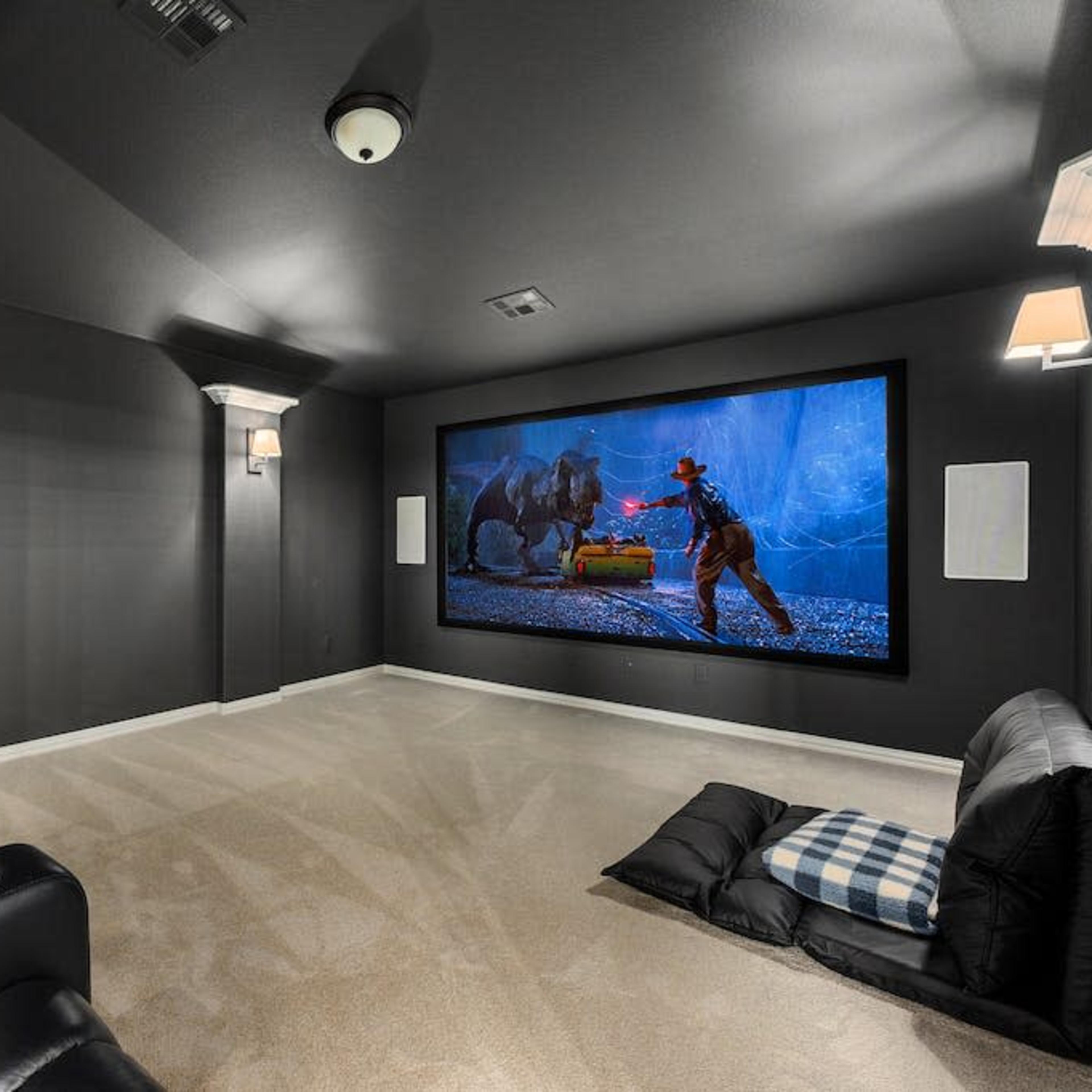 A home theater room featuring black reclining chairs, carpeted flooring, and a large projection screen displaying a scene of a cowboy confronting a bear.