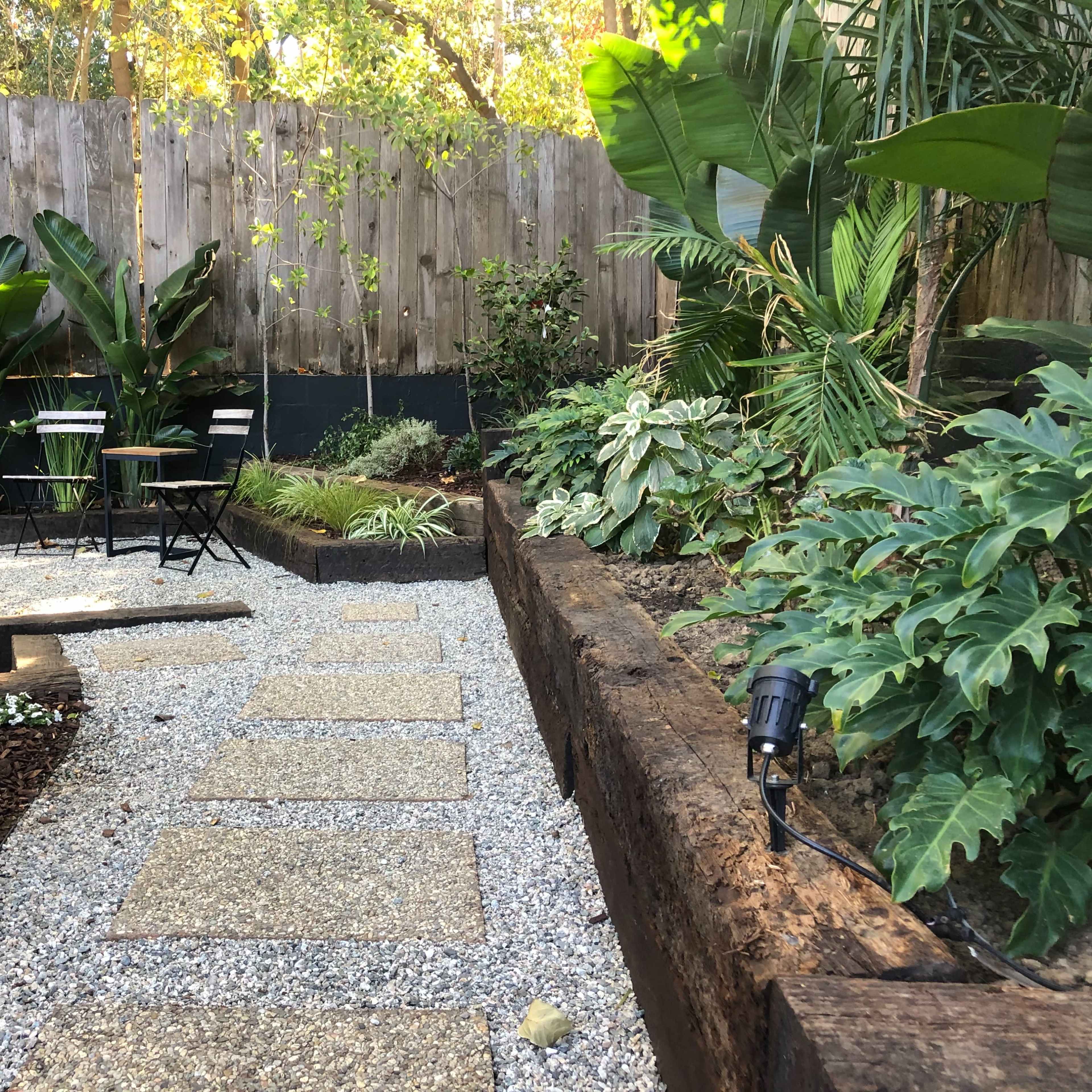 A landscaped garden path made of gravel and stone slabs, bordered by greenery and shaded by tall plants, with a seating area and a fountain at one side.