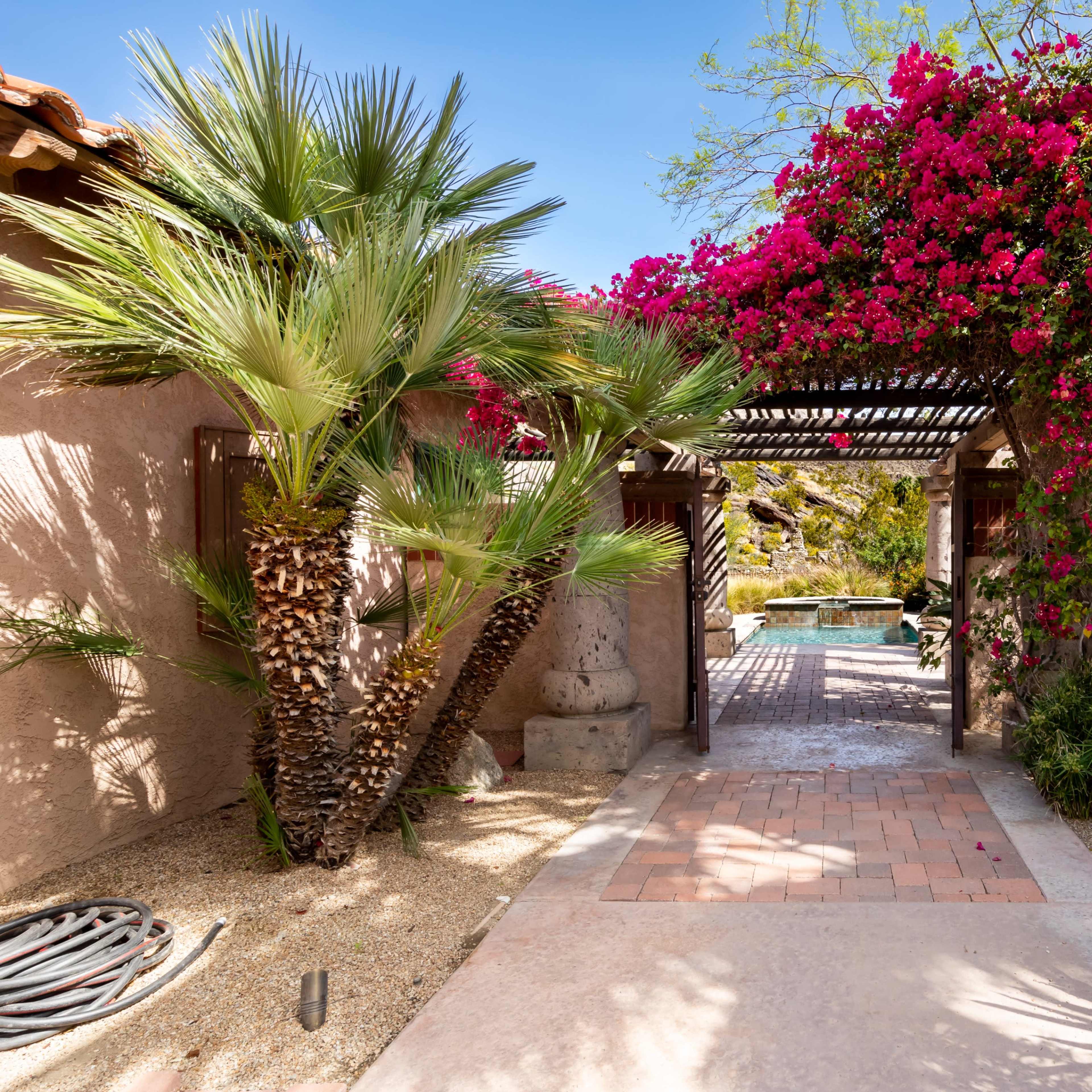 A stone pathway leads through a flower-covered archway to a pool area surrounded by lush landscaping.