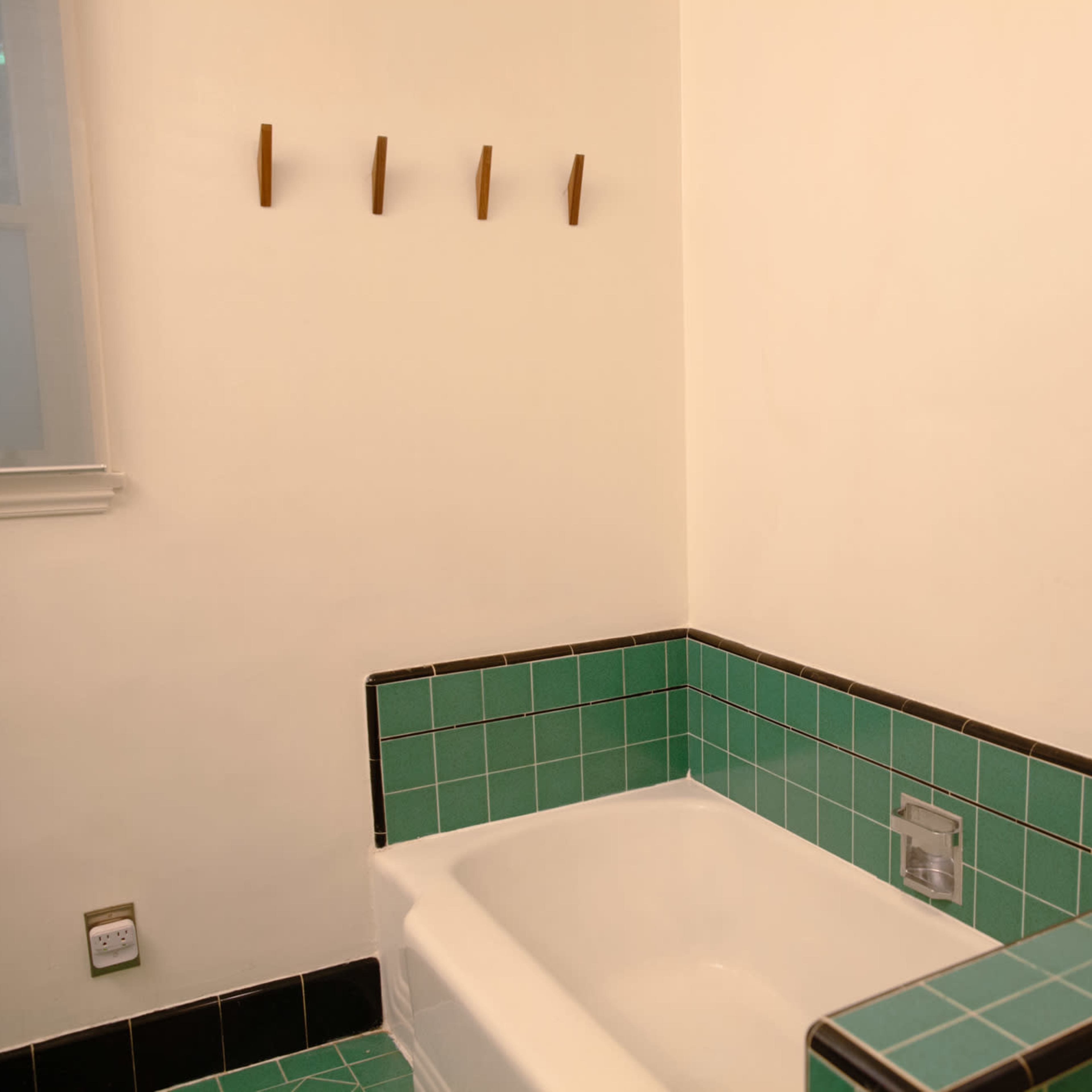 A bathroom with a green tiled bathtub, a small plant in a woven pot on the edge, and wall hooks in the background.