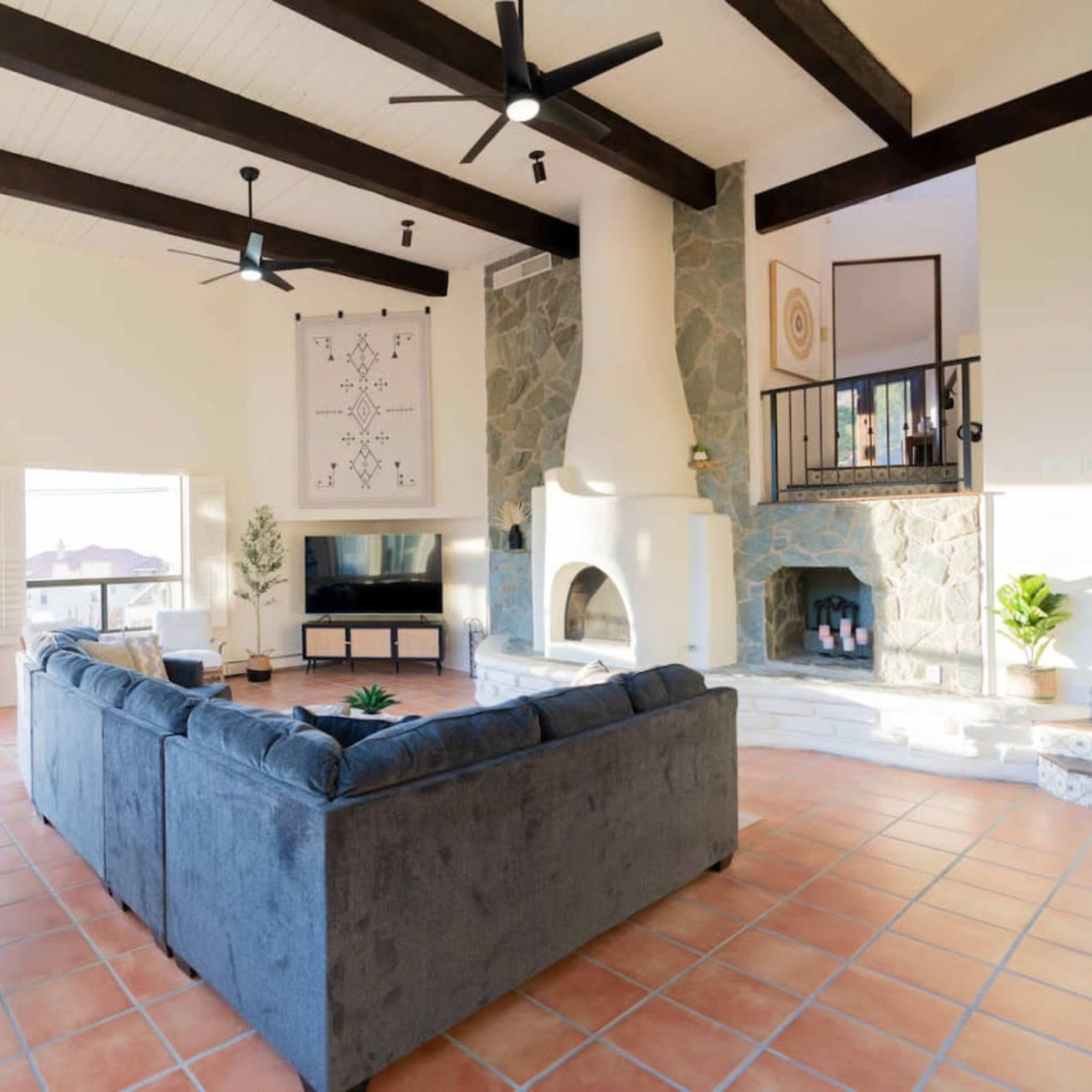 A spacious living room with a high ceiling, featuring a stone fireplace, large windows, and a sectional sofa arranged around a central area.