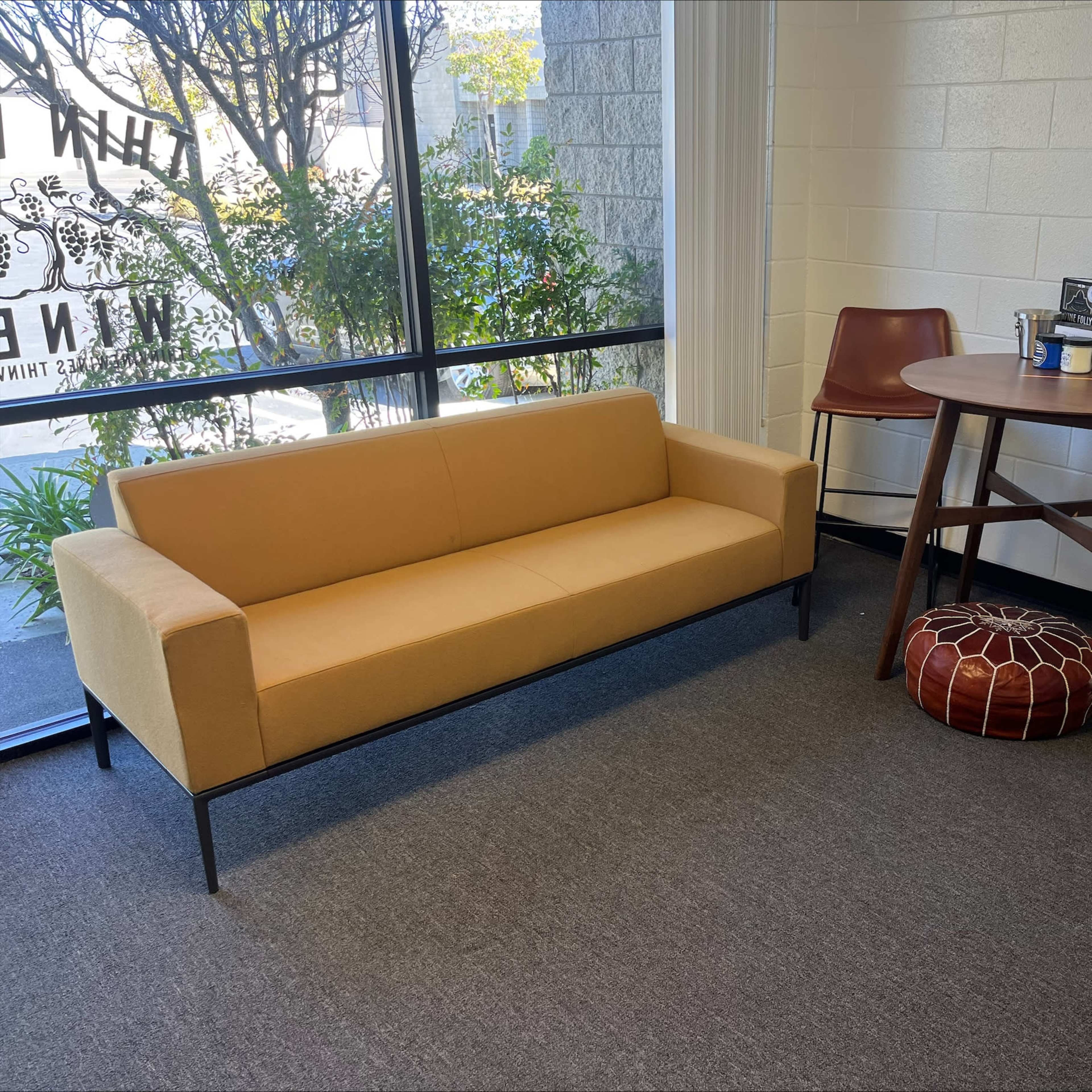 A small seating area with a beige sofa, two brown bar stools, a round wooden table, and a leather pouf, situated next to a large window with greenery outside.