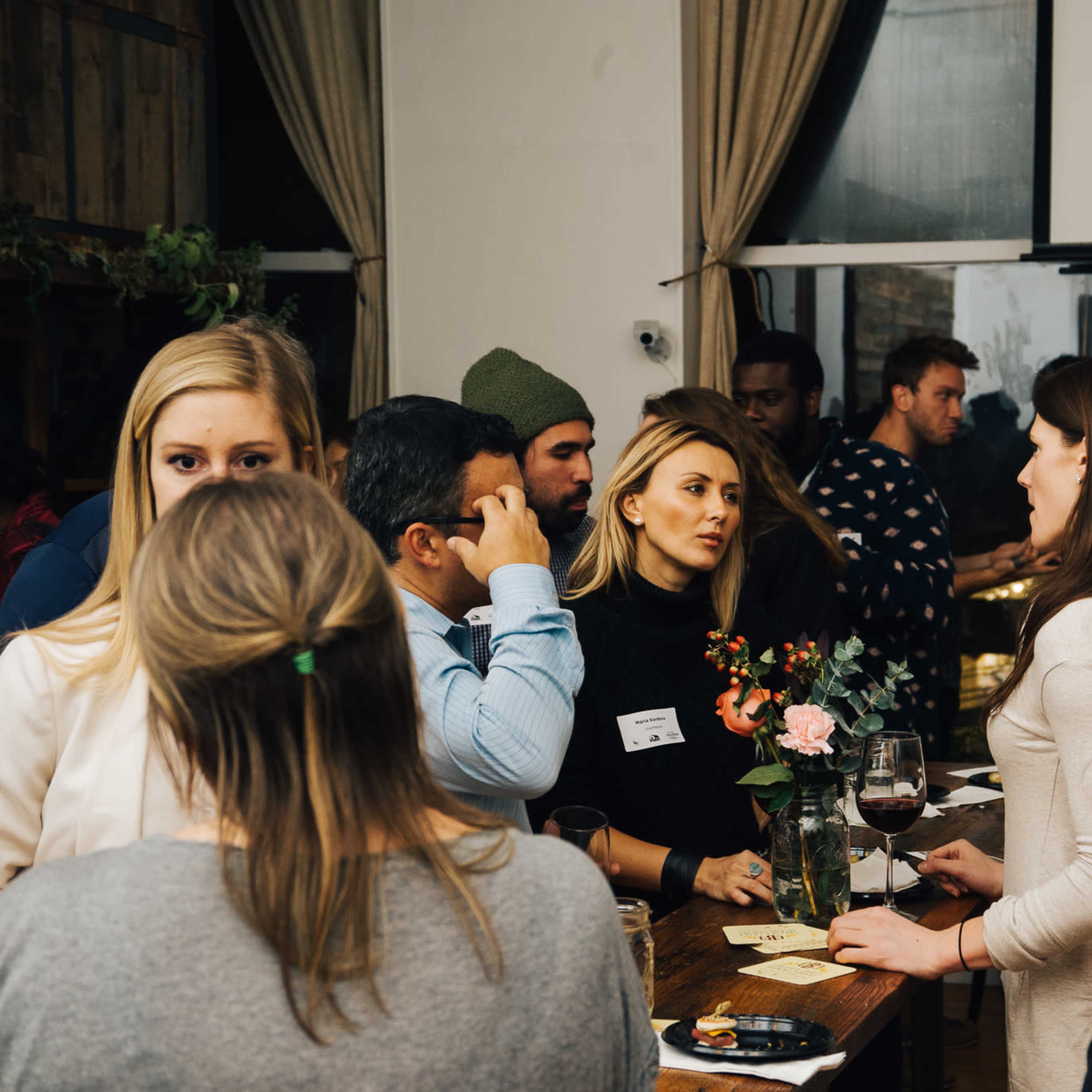 A group of people converses at a table in a social gathering with drinks and flowers.