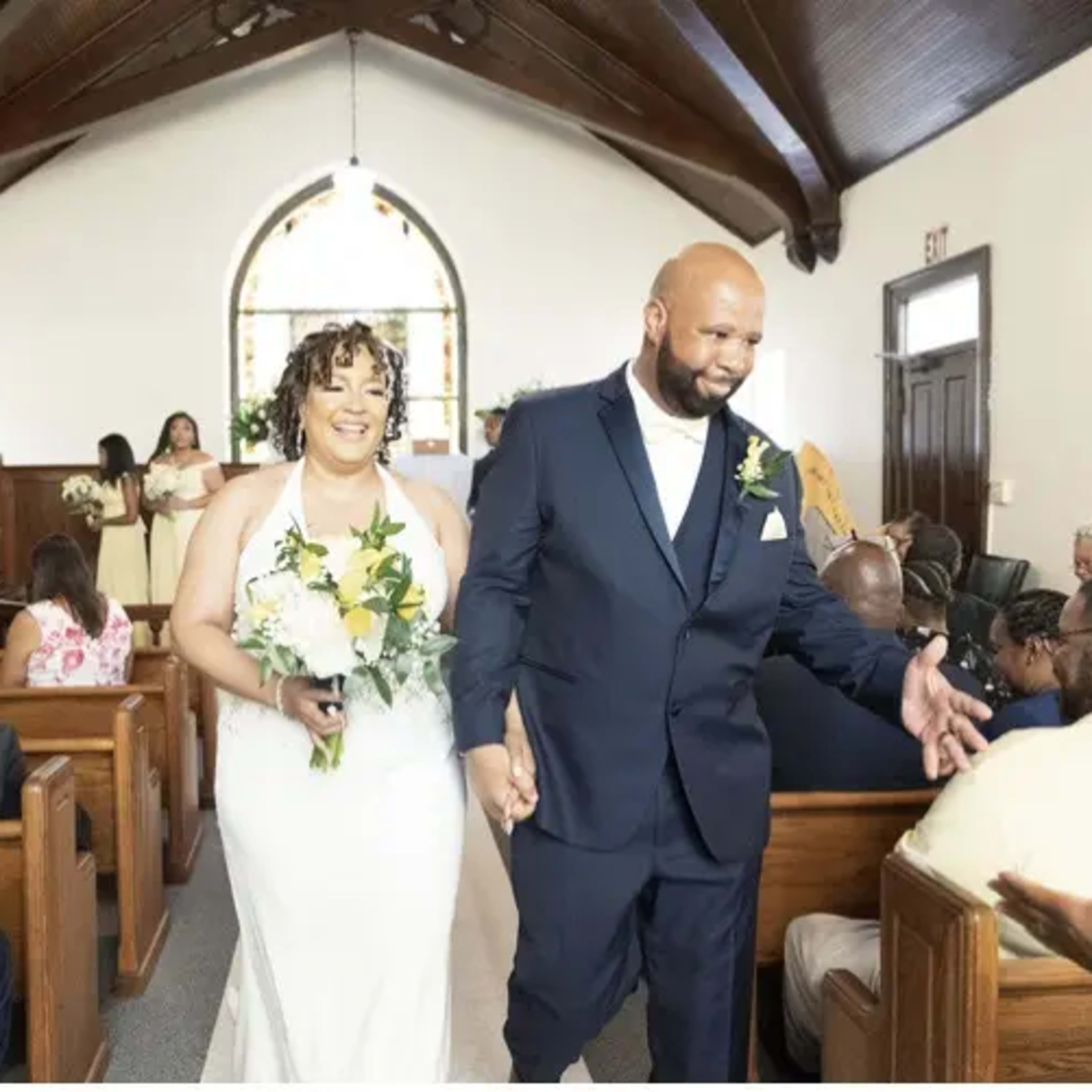 A bride and groom walk down the aisle of a church while guests seated in wooden pews smile and congratulate them.