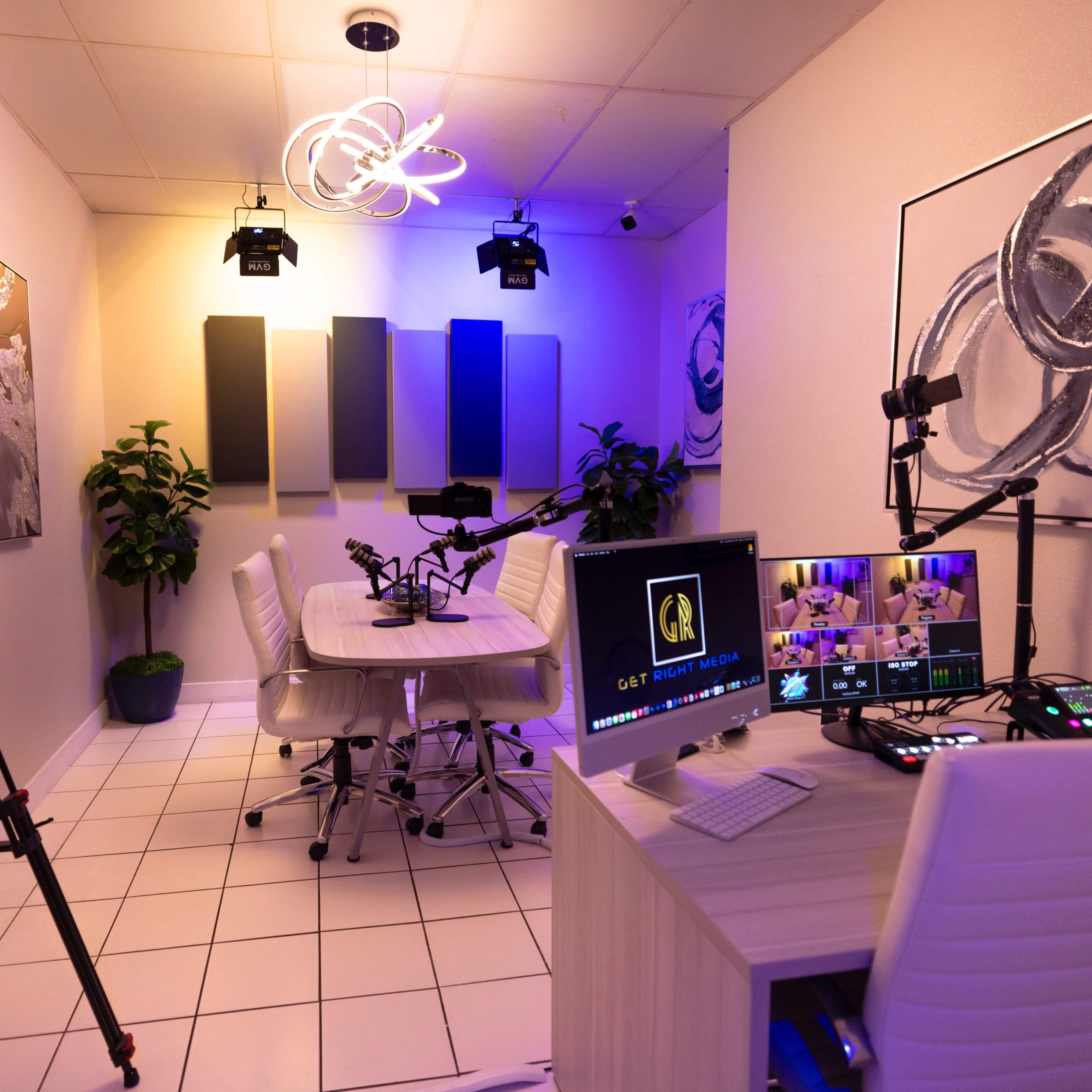A recording studio with a round table, multiple cameras on tripods, and a computer setup displaying a live stream interface, illuminated by overhead lights.