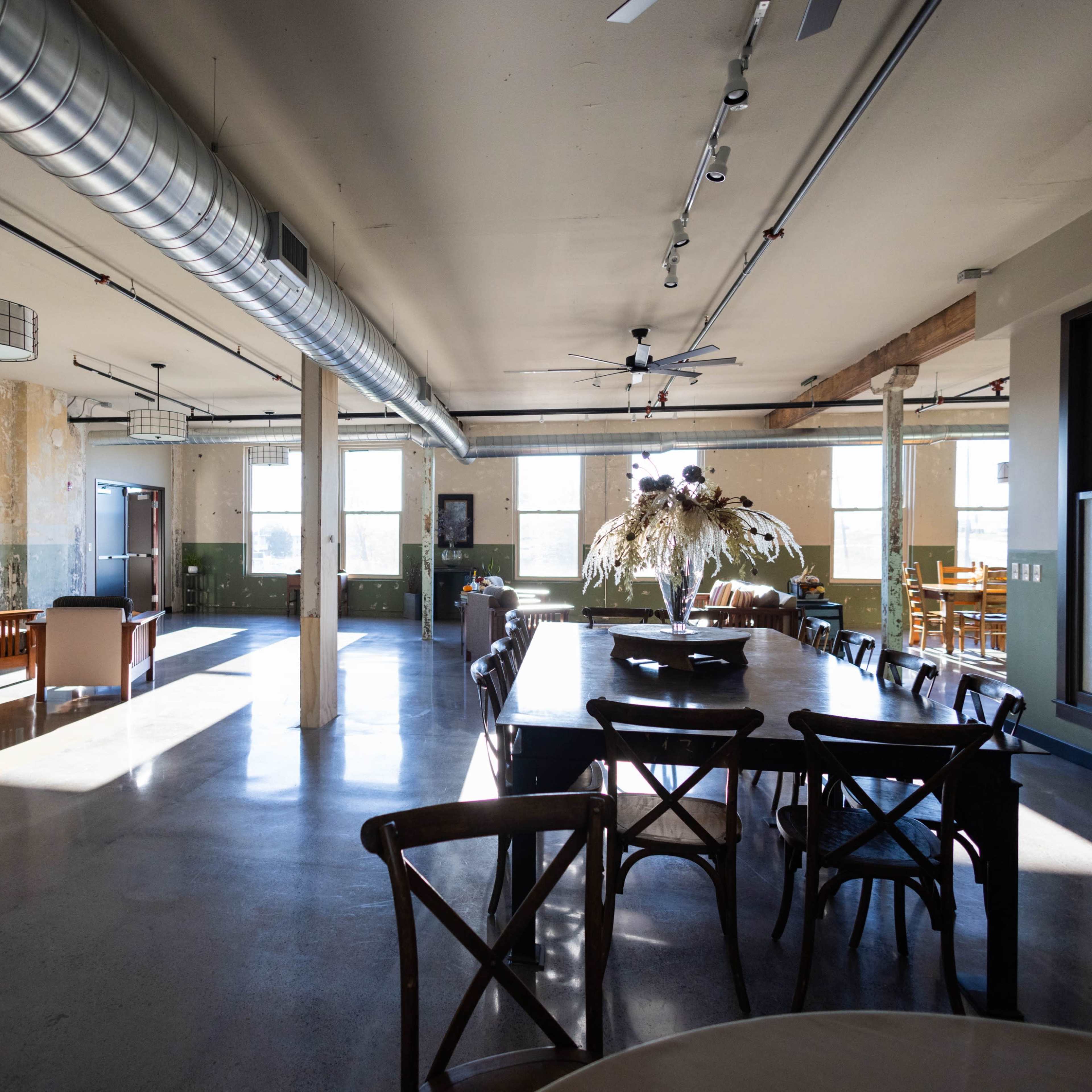 An airy, industrial-style interior space with large windows, a long dining table, and a mix of seating areas.