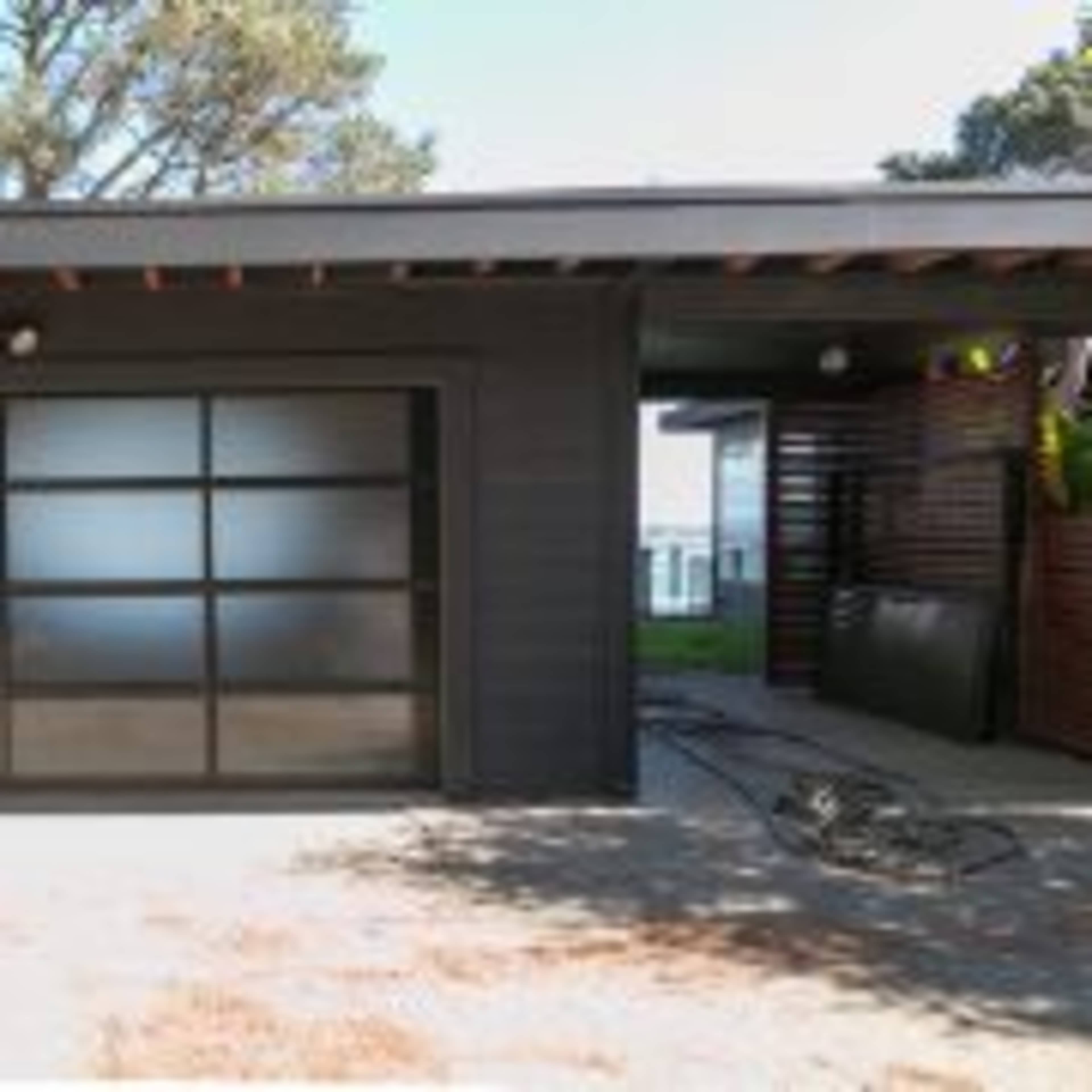 A modern garage with a large glass door, adjacent to a covered entrance of a house, surrounded by trees.