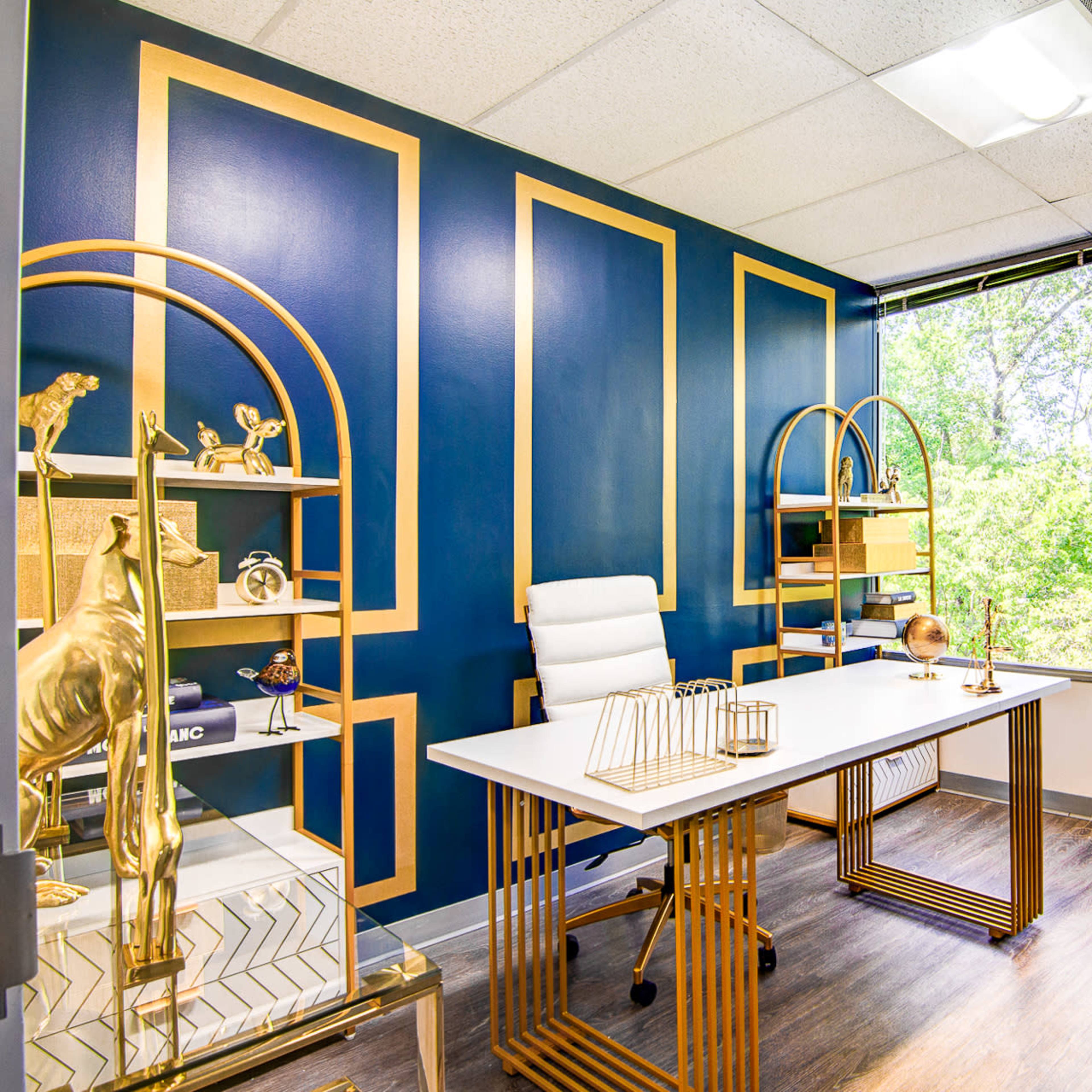 A modern office space features a desk with a white chair, gold accents, and large windows overlooking greenery.