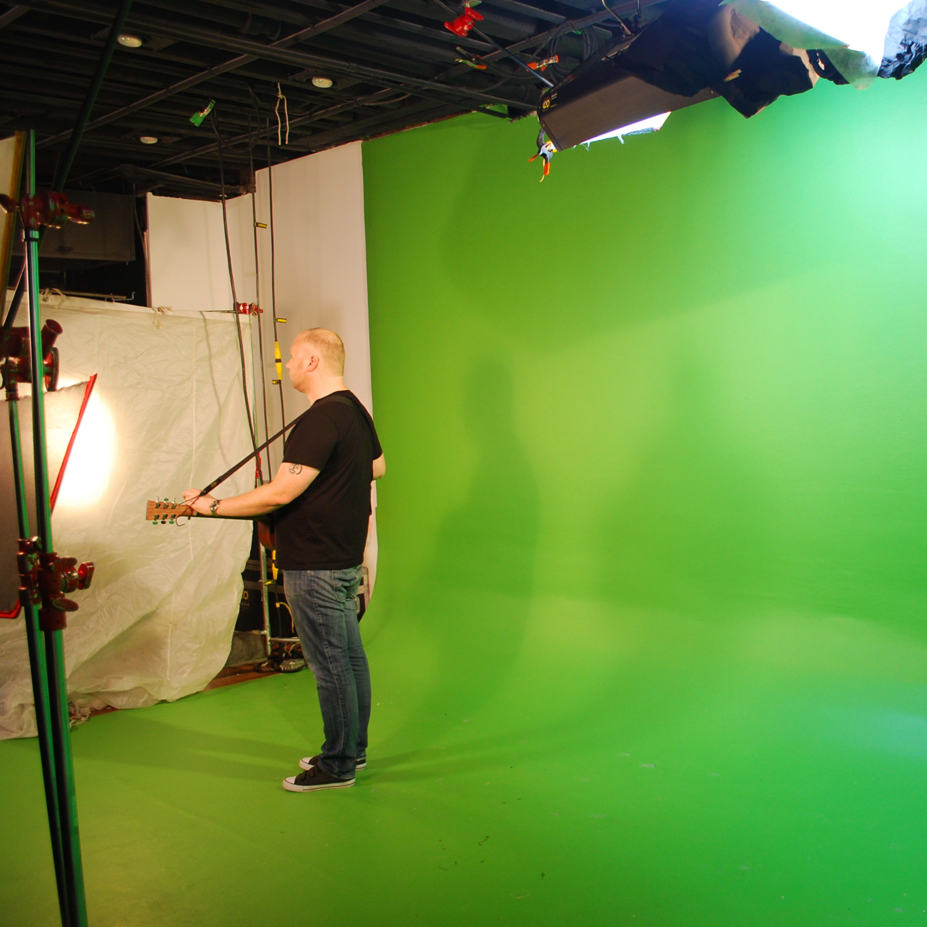 A person stands with a guitar in front of a green screen, with studio lights illuminating the scene.