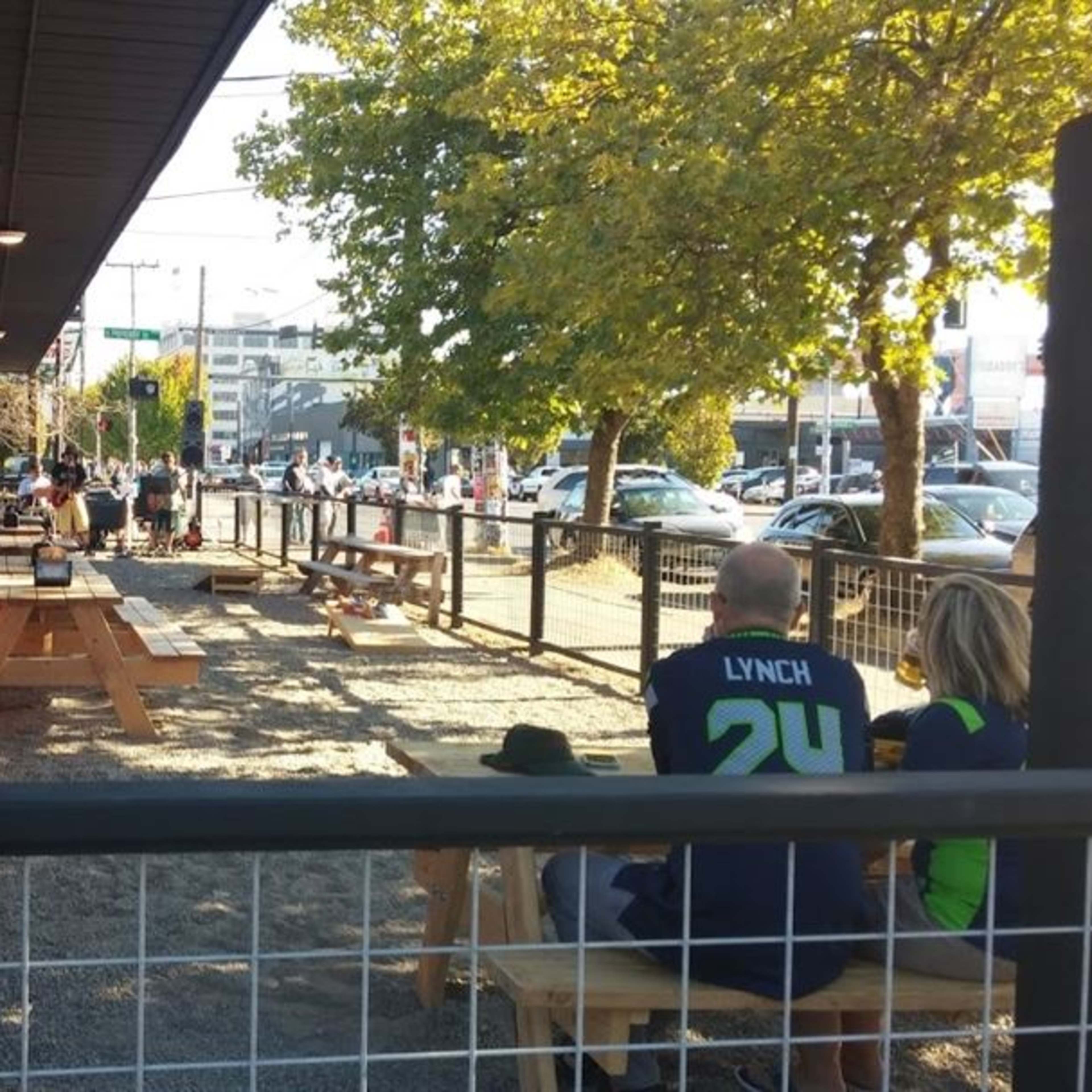 A patio area with wooden picnic tables is filled with patrons, and a couple sits together, while cars are parked along the street nearby.