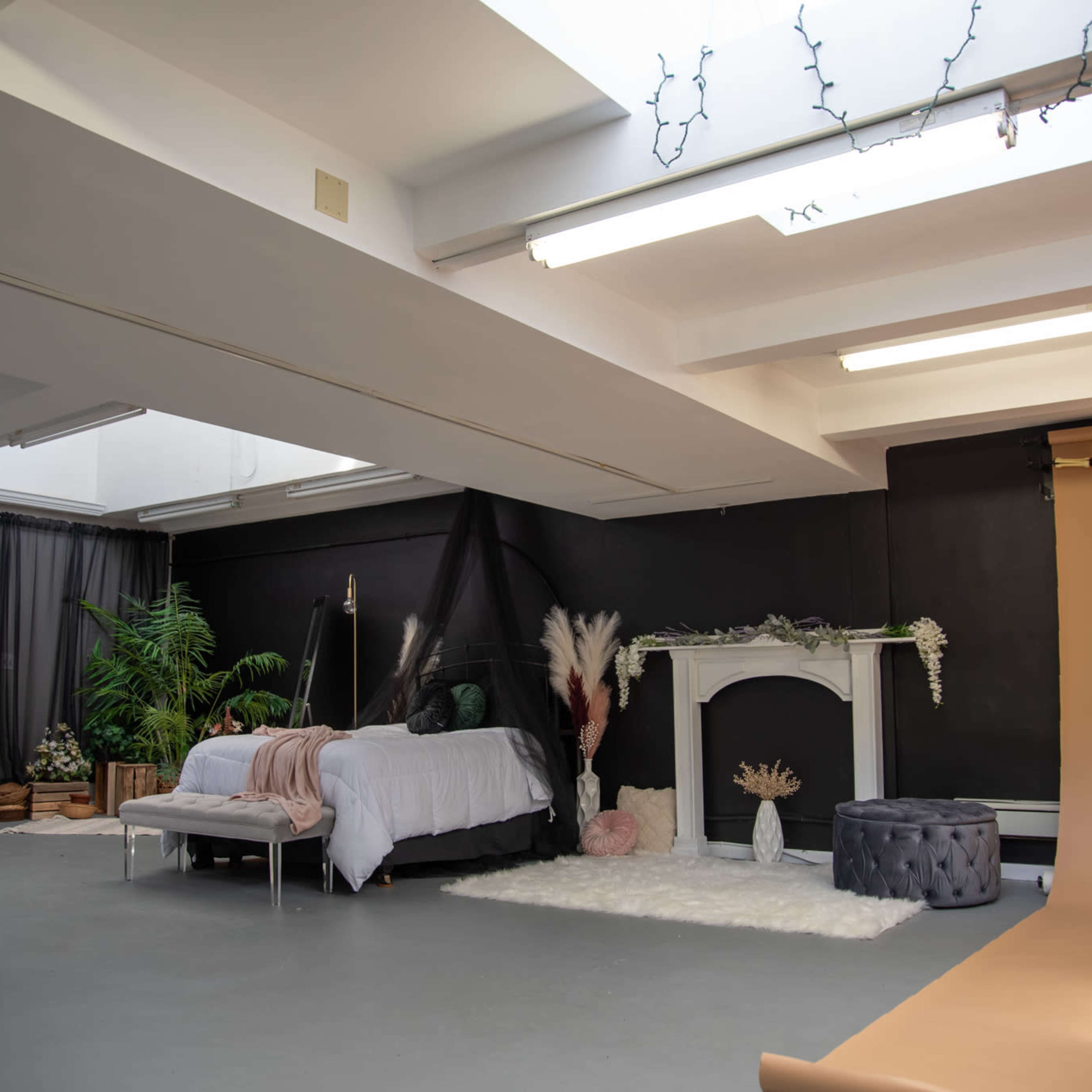 A spacious studio setting with a bed, a decorative fireplace, plants, and a neutral-toned backdrop.
