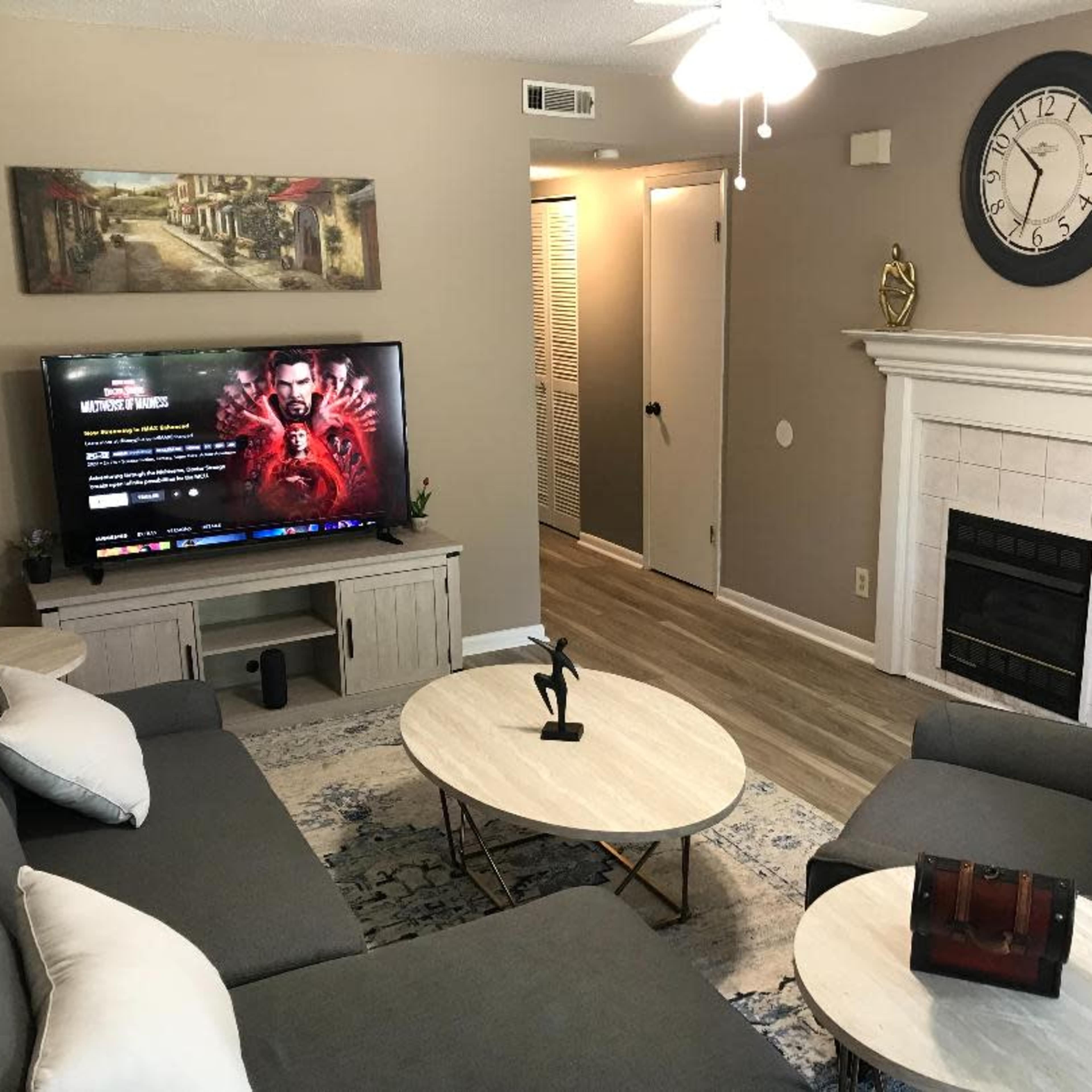 The living room features a television on a stand, a coffee table, two sofas, and a fireplace, with a painting on the wall and a clock above the mantel.