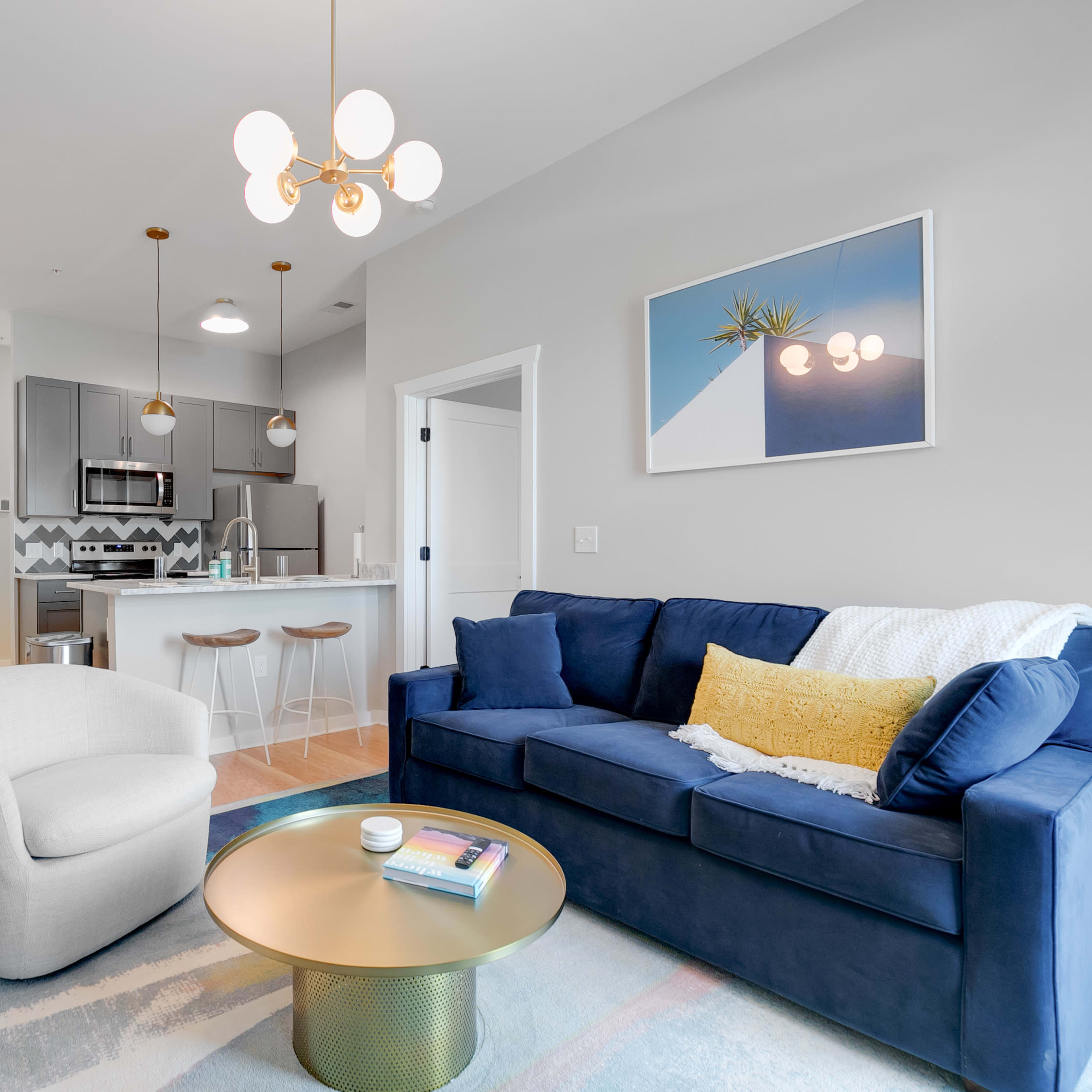 A modern living room with a blue sofa, a white armchair, and a gold coffee table, adjacent to a kitchen area with gray cabinets and stainless steel appliances.