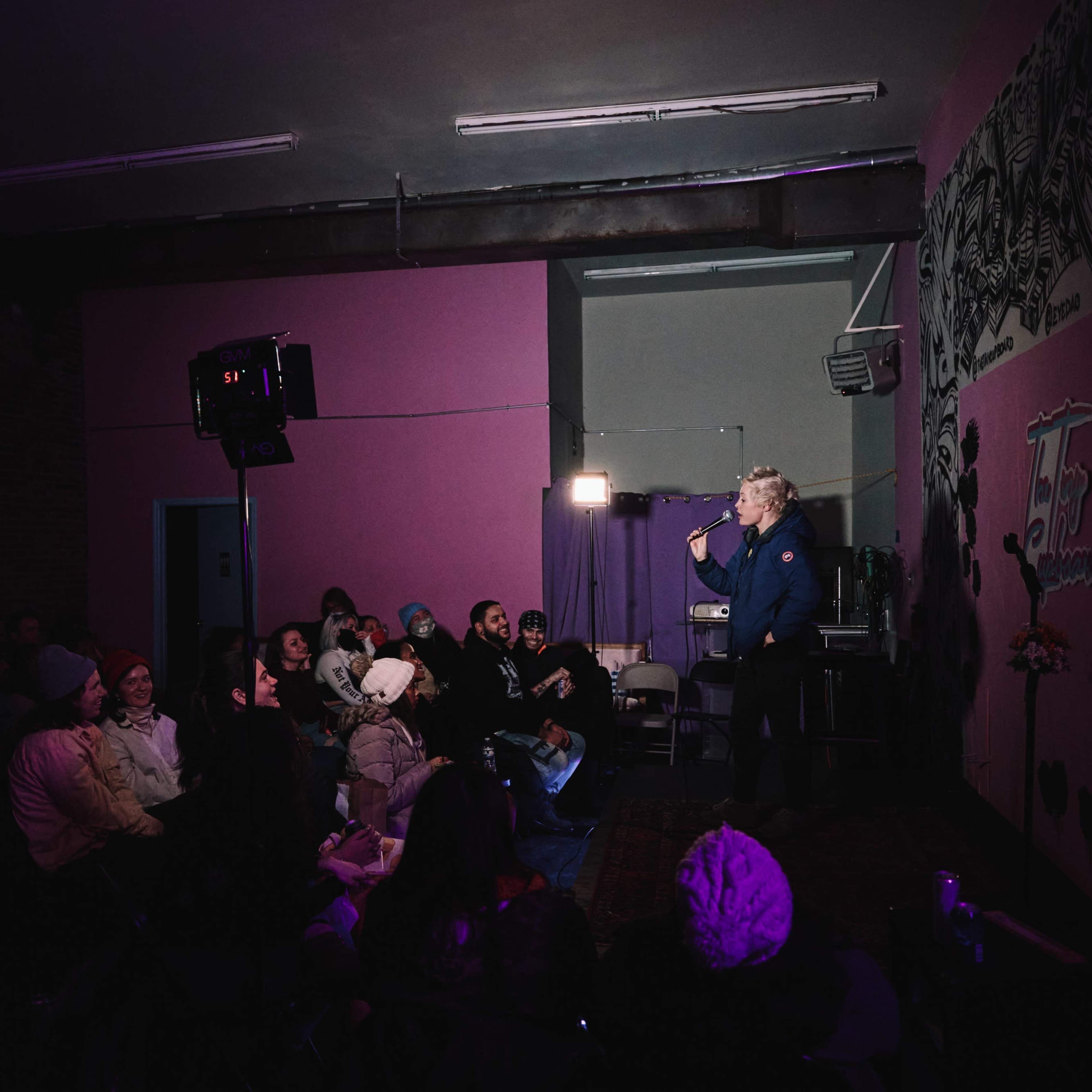 A performer stands on stage in front of an audience seated in a dimly lit room with colorful walls.