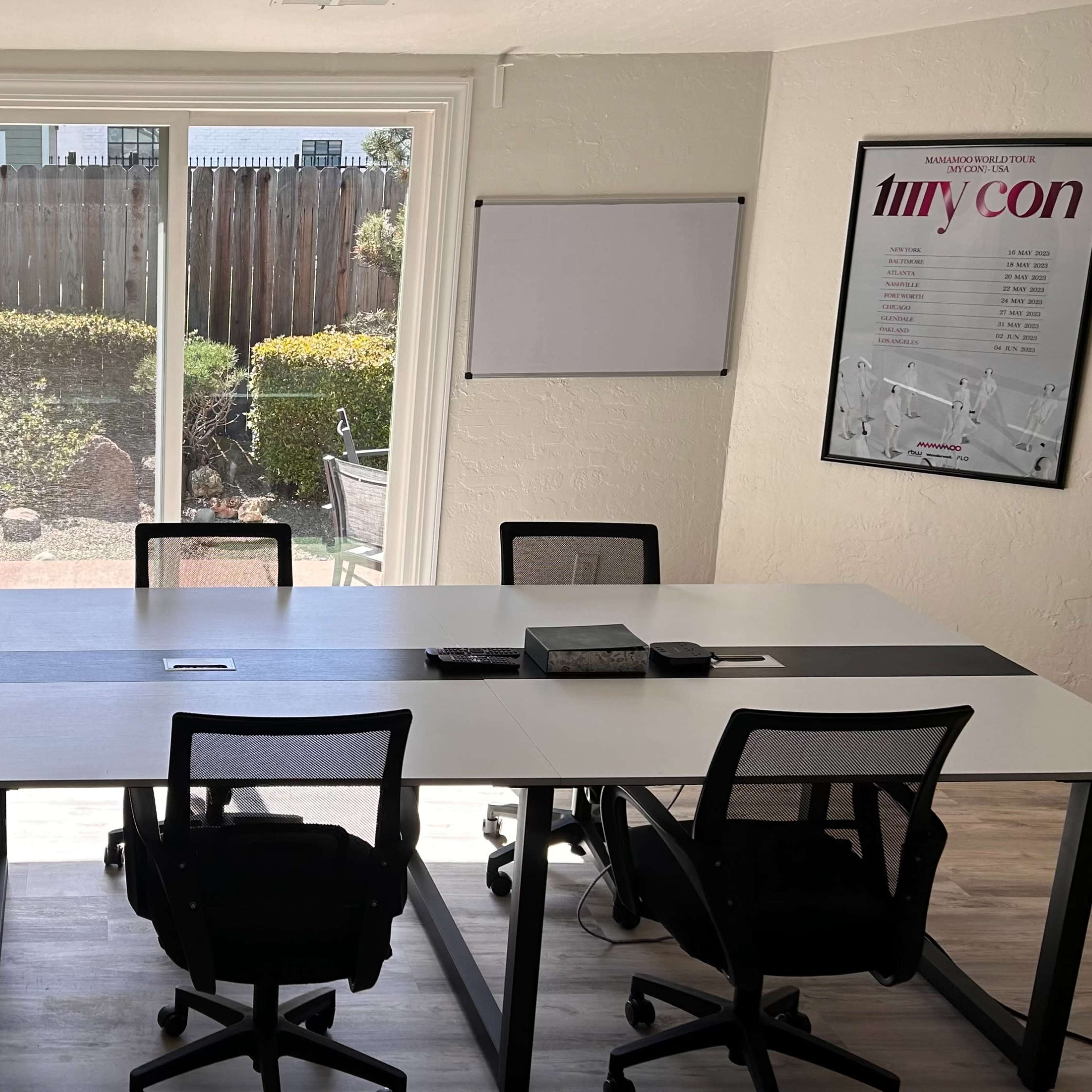 A bright meeting room features a large table surrounded by black chairs, with windows overlooking a garden and posters on the walls.