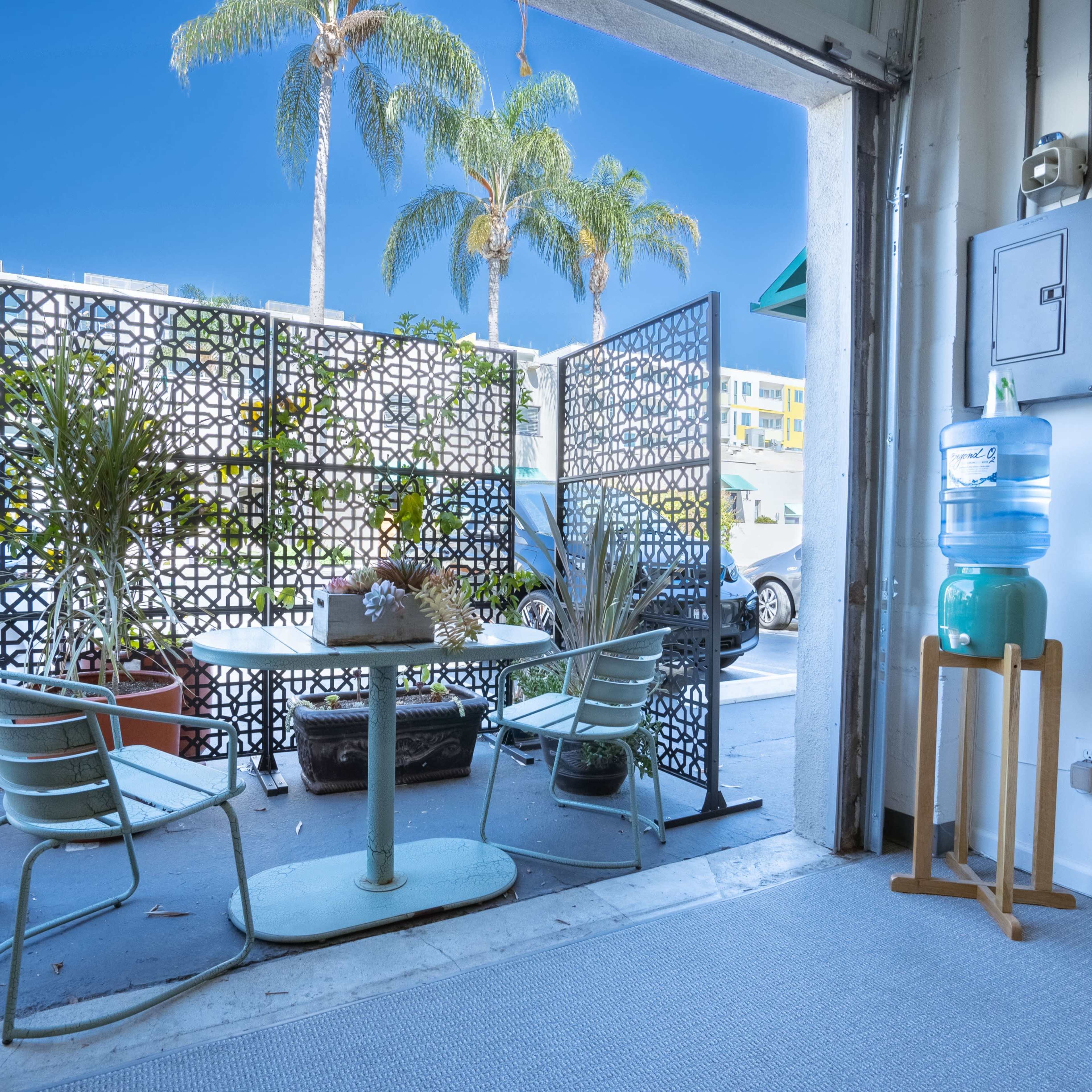 A table and chairs are set up near an open garage door, with a decorative screen and a water cooler nearby, and palm trees visible outside.