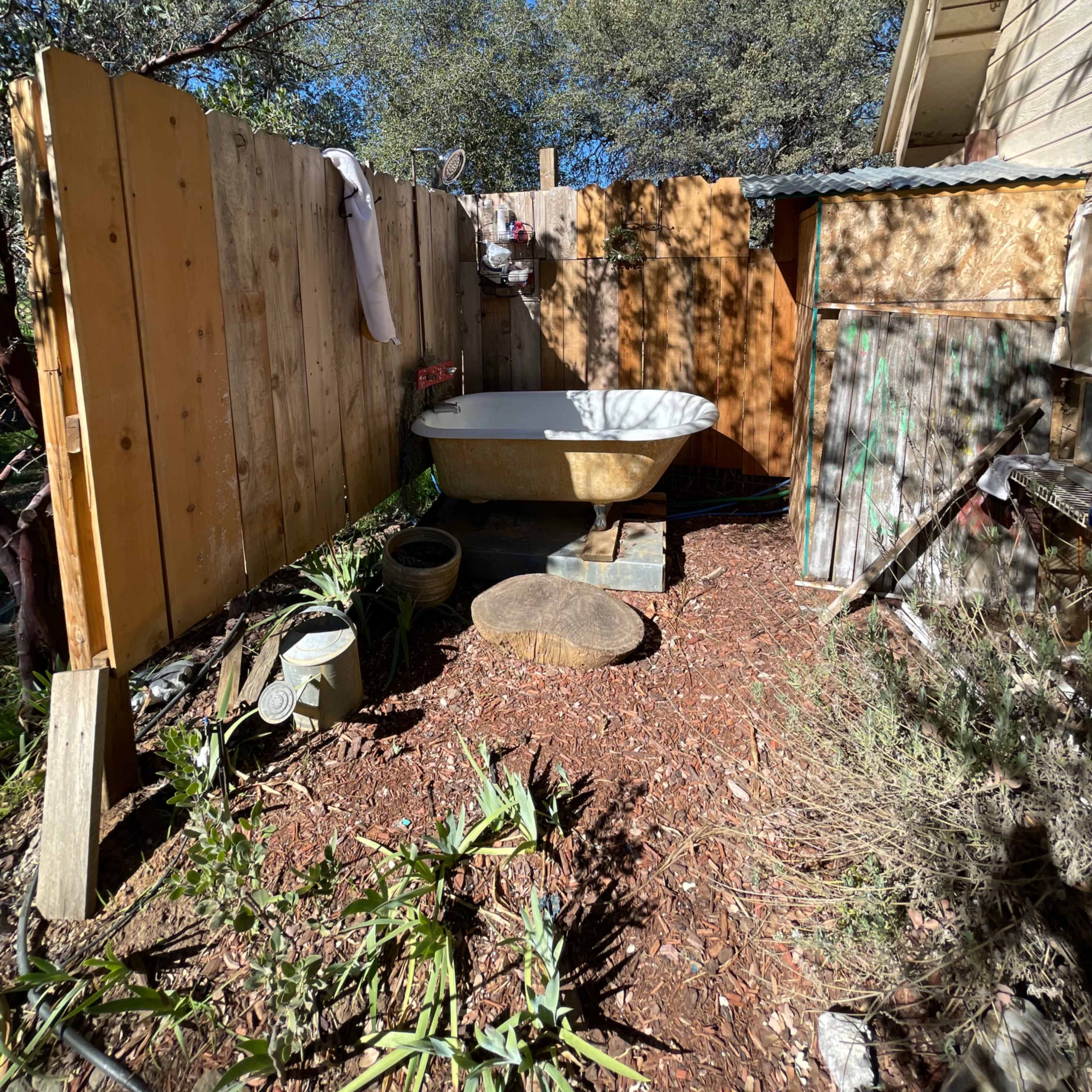 An outdoor area features a freestanding bathtub surrounded by wooden fencing and various plants.