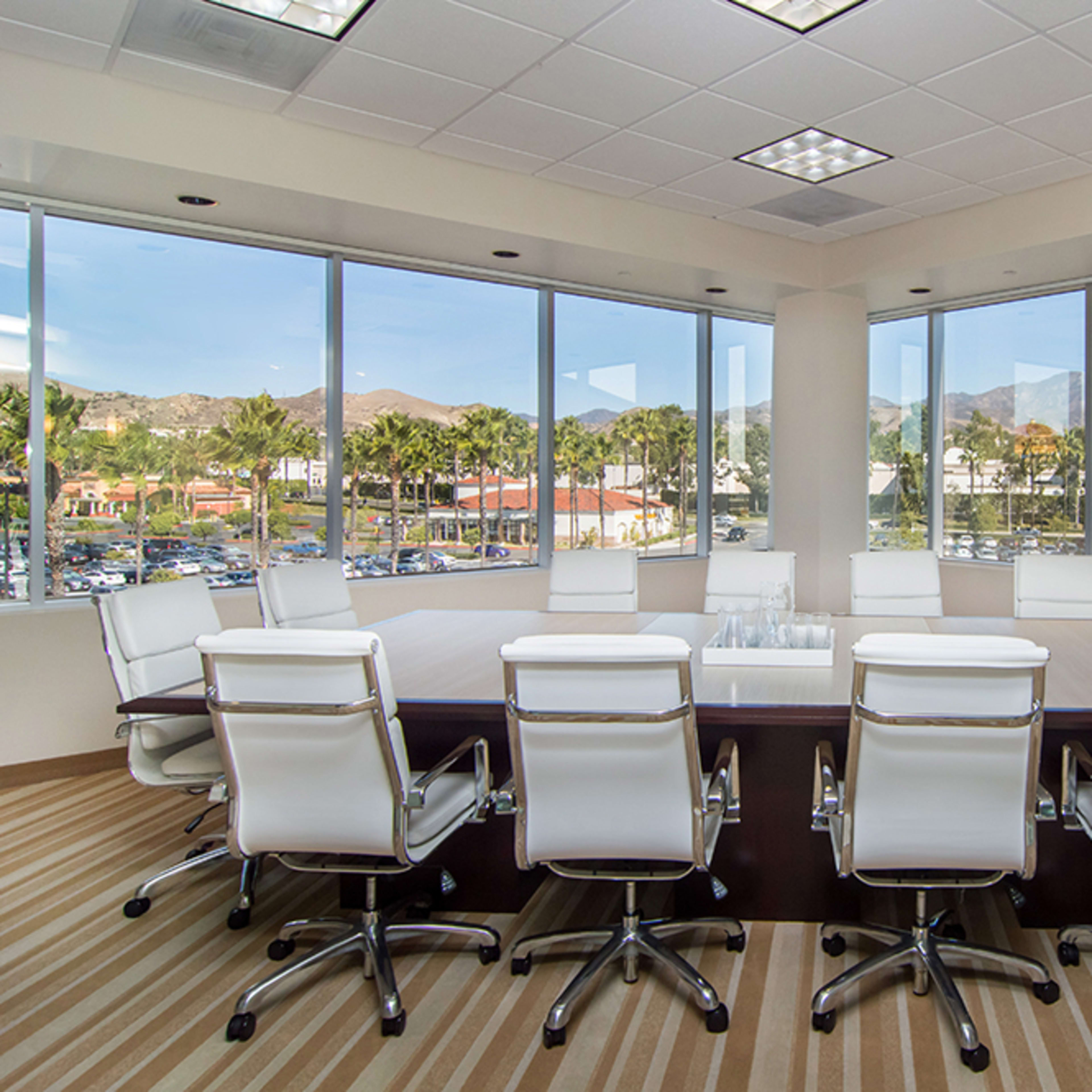 A conference room with a large table surrounded by white chairs and windows offering a view of mountains and palm trees outside.