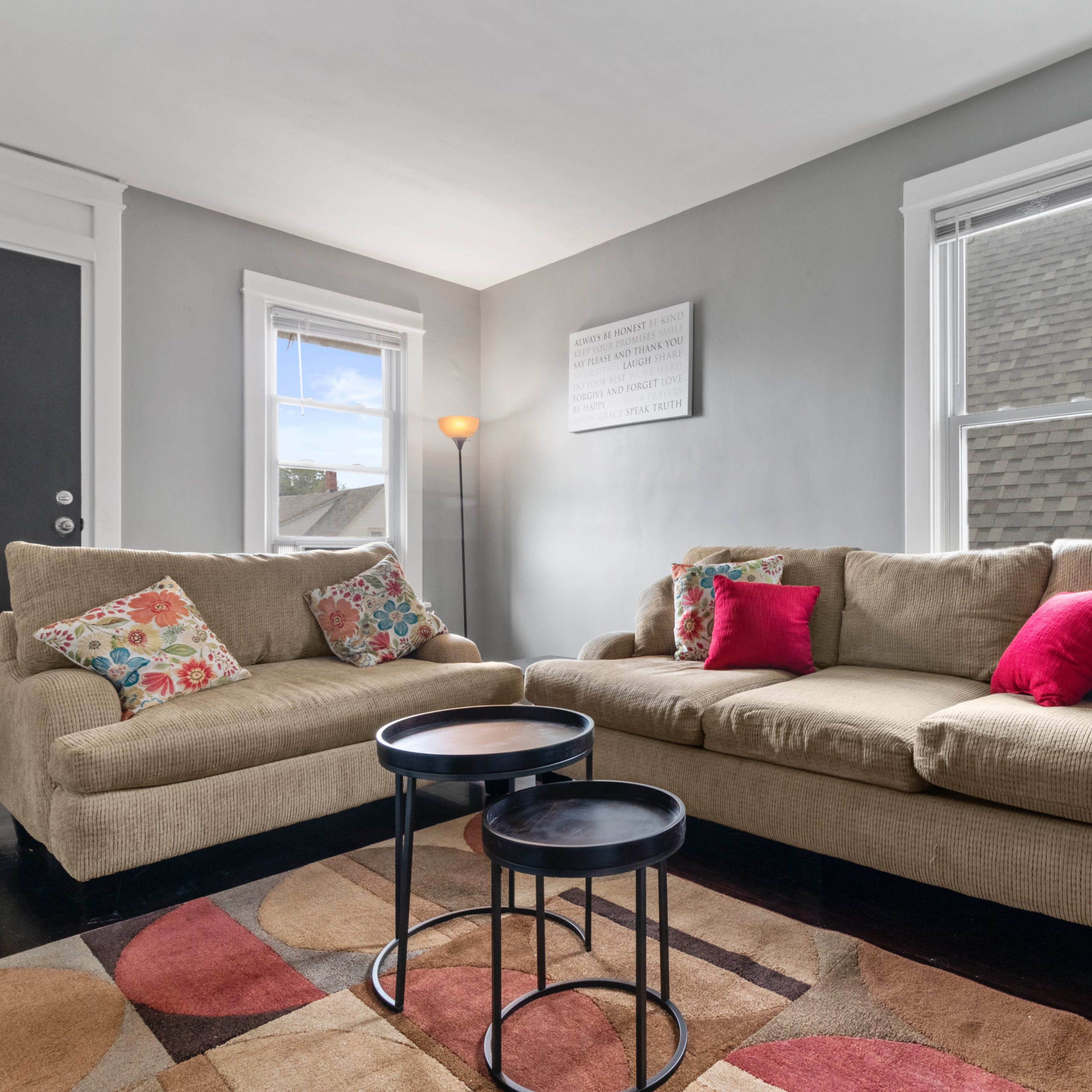 A cozy living room featuring a beige sofa with colorful cushions, two small black tables, and large windows allowing natural light.
