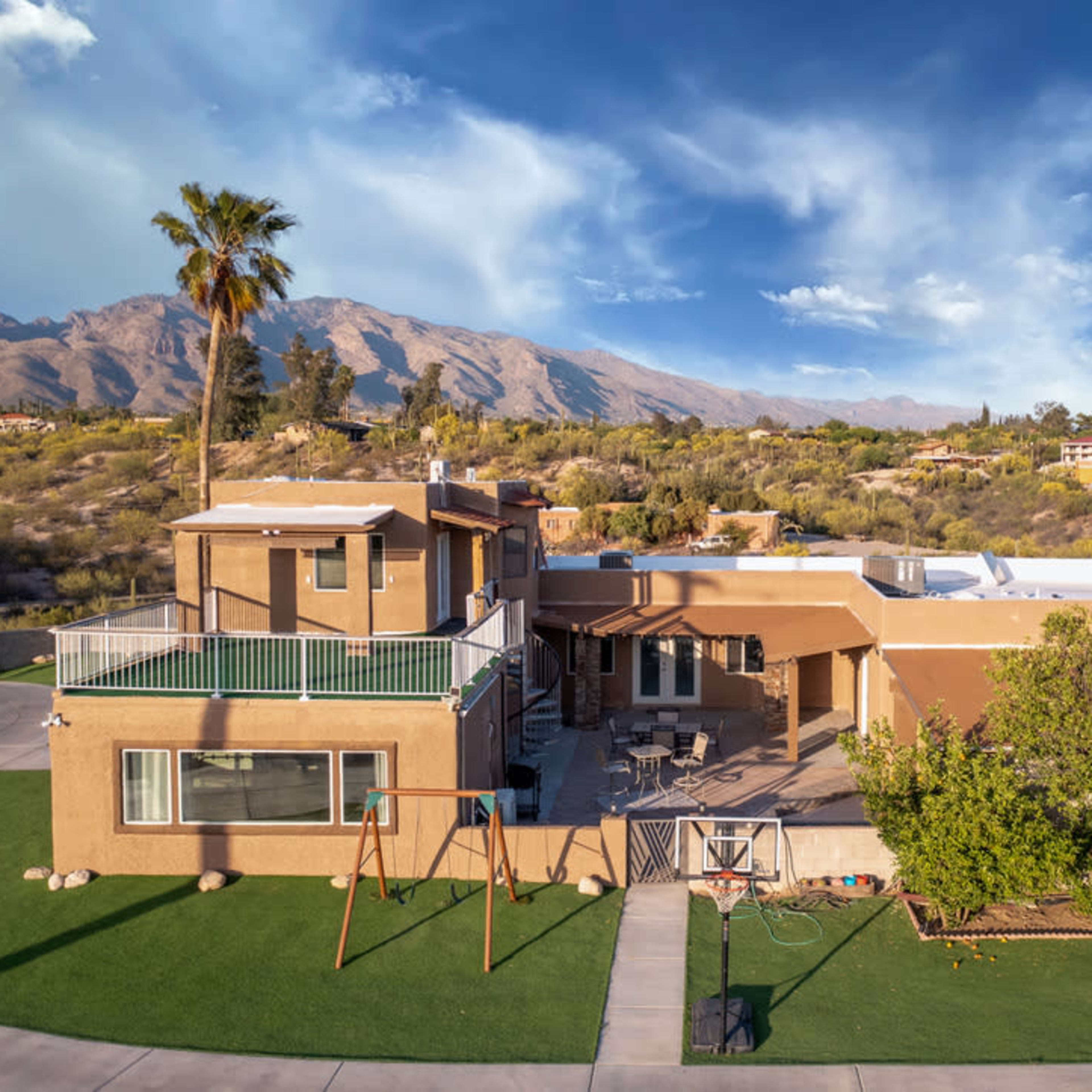 A large tan house with a patio and a swing set in the front yard, surrounded by mountains and palm trees.