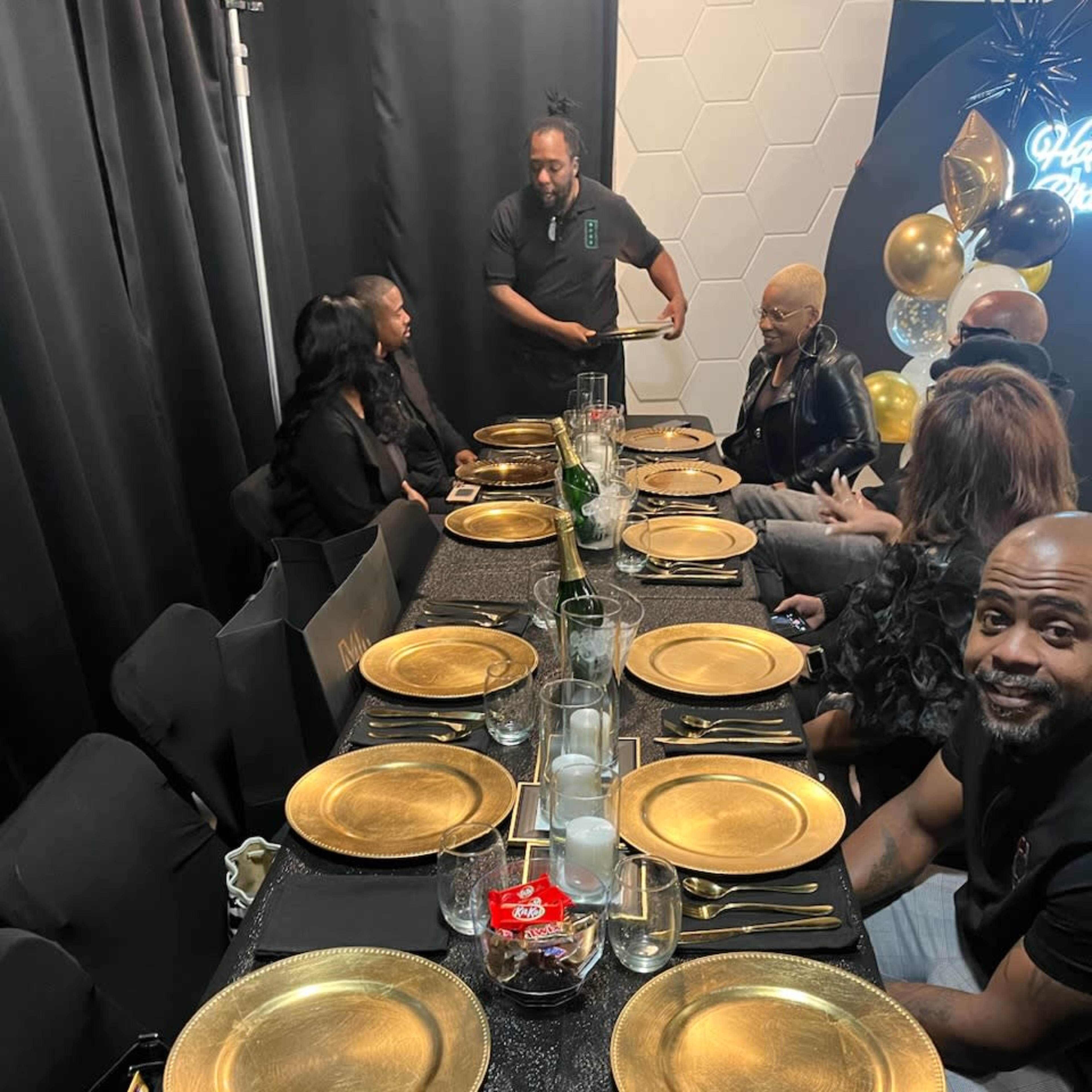 A elegantly set dining table with gold plates and silverware is prepared for a gathering, while a person interacts with seated guests.