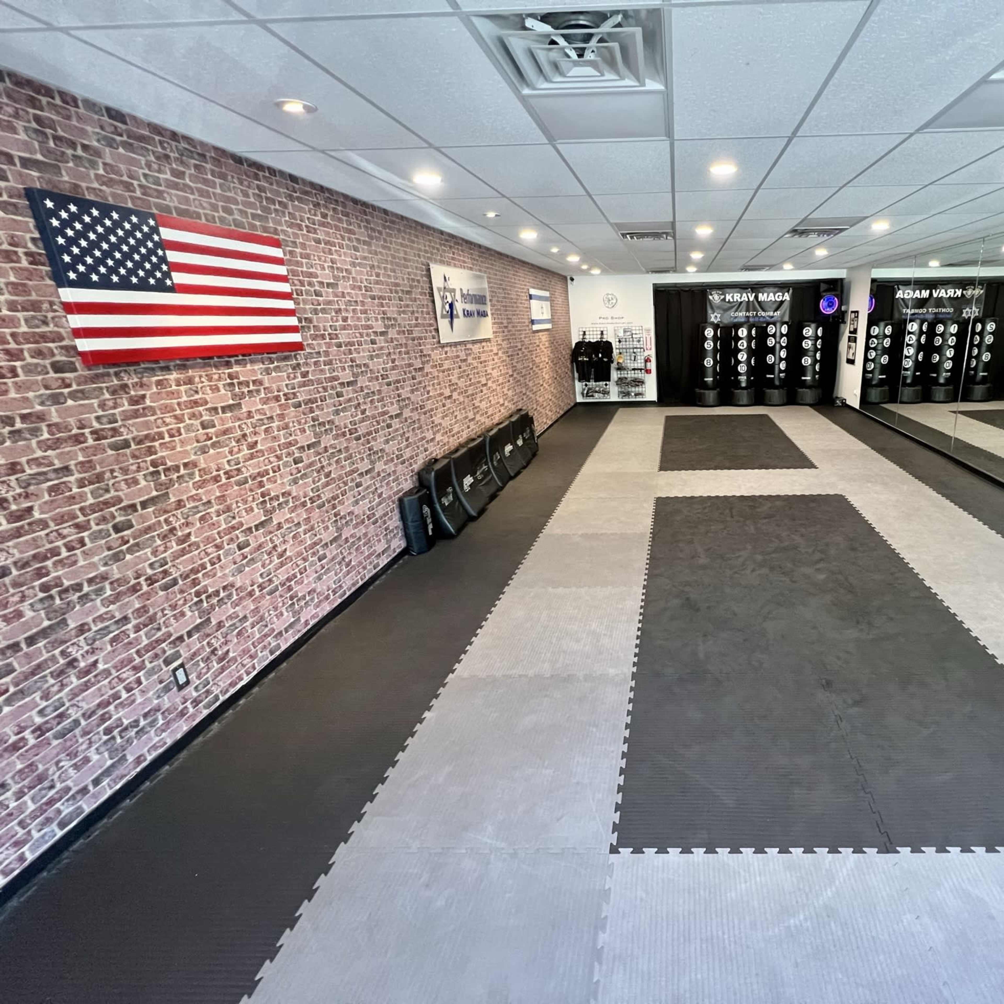 A spacious martial arts studio with a brick wall, an American flag, and mats on the floor, along with various training equipment displayed.