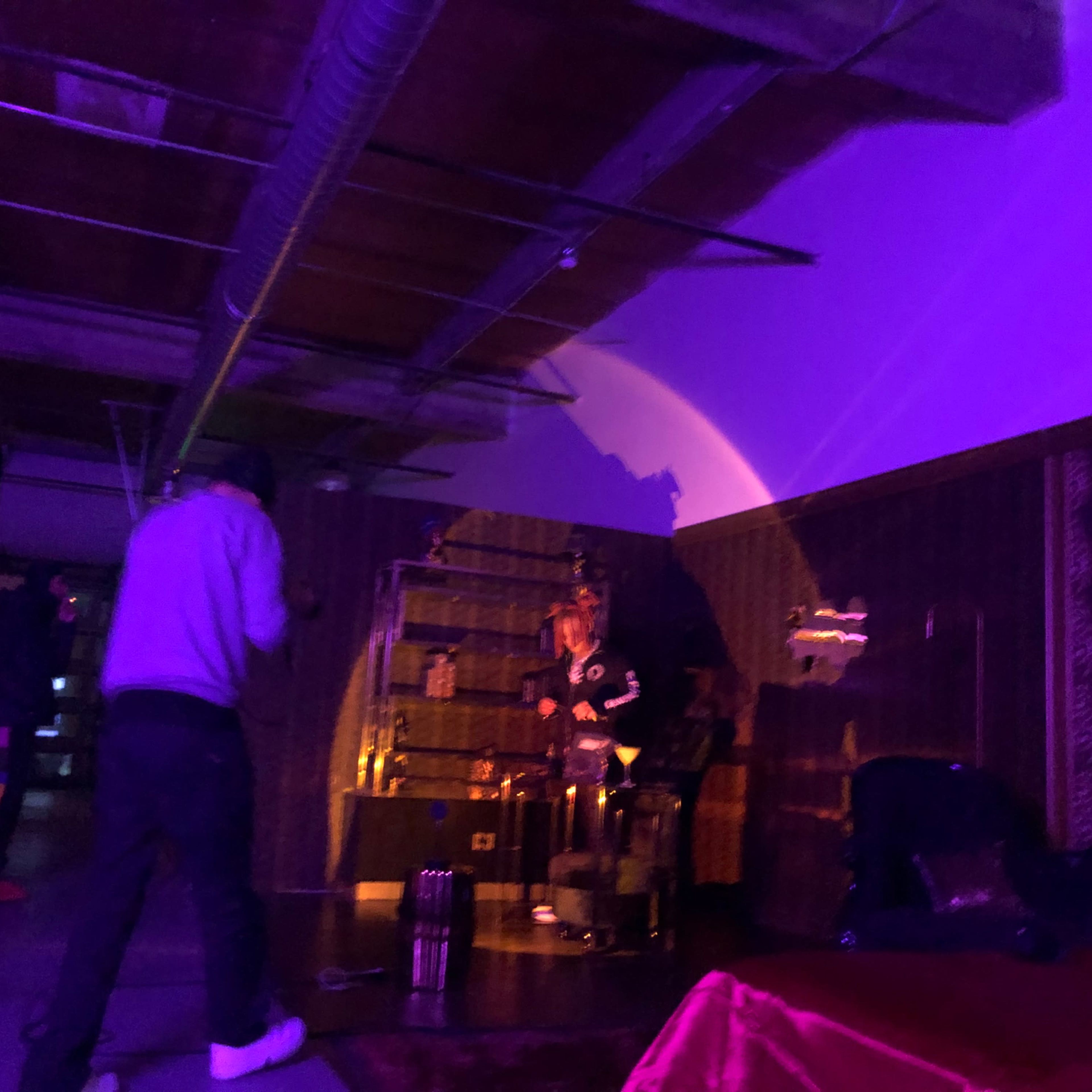 A person stands in a dimly lit room with purple lighting, interacting with a woman seated near a shelf filled with objects.