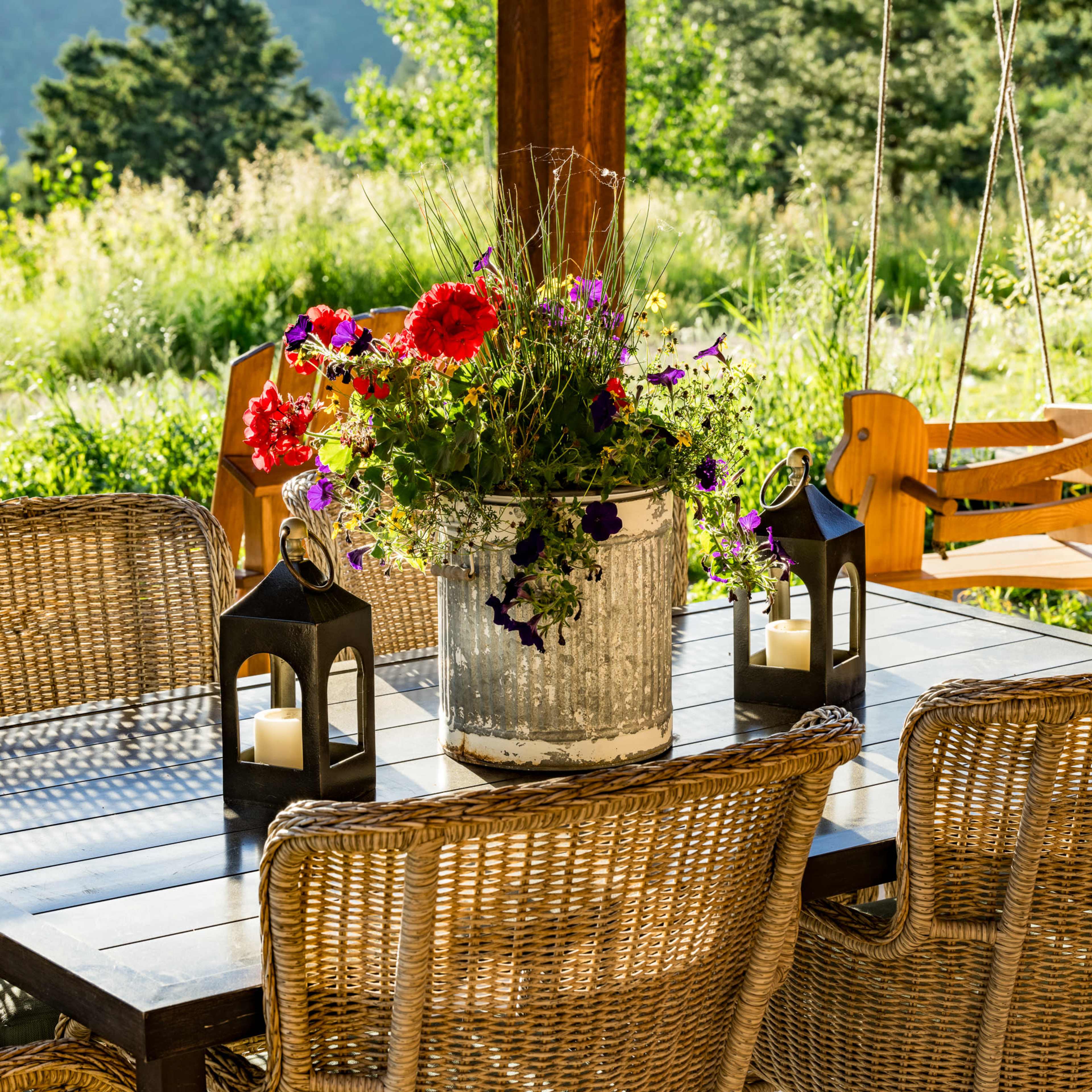 A rustic outdoor dining area features a wooden table surrounded by wicker chairs, adorned with a large floral arrangement in a metal container and two decorative lanterns.