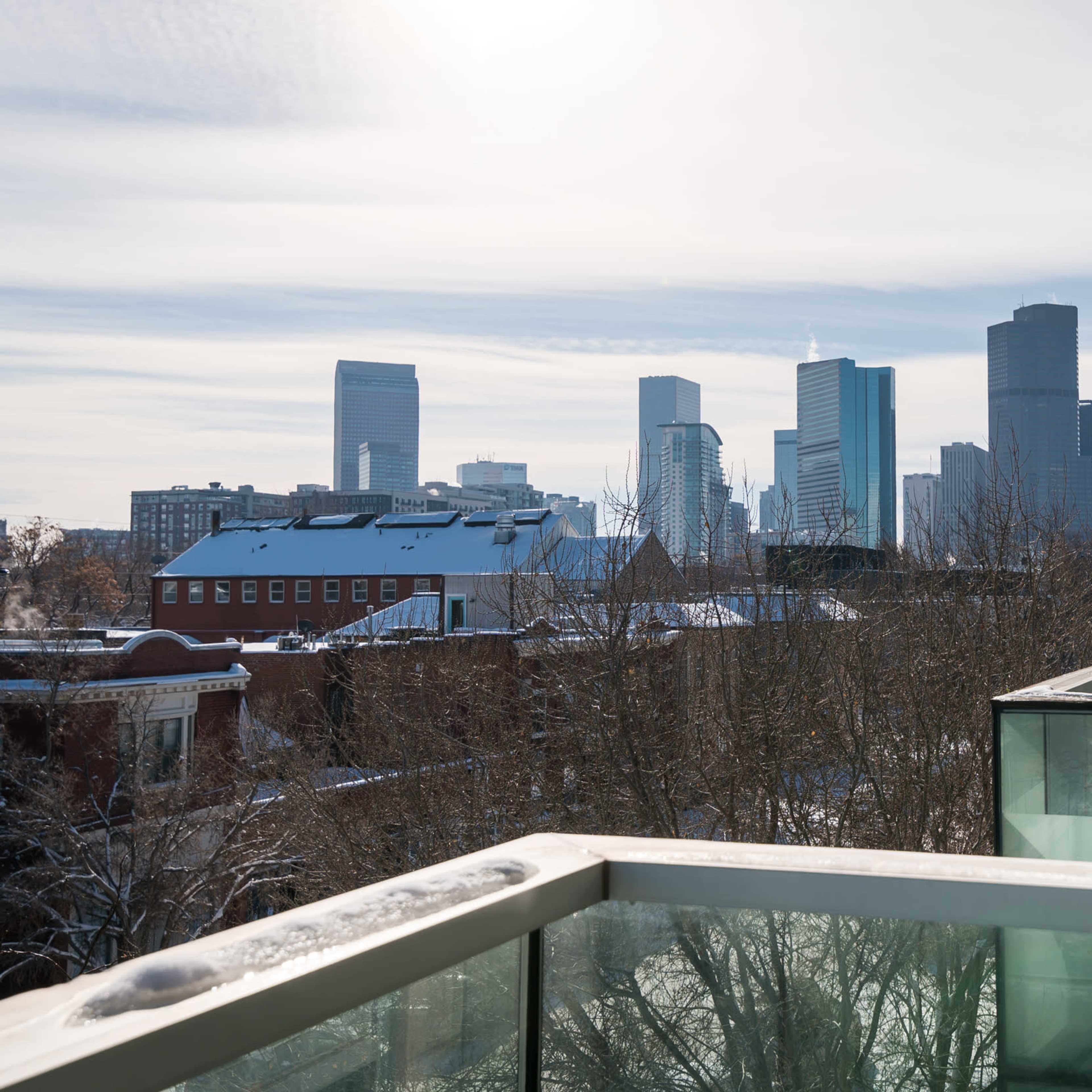 A view of a city skyline with several modern skyscrapers in the background, framed by bare trees and a balcony in the foreground.