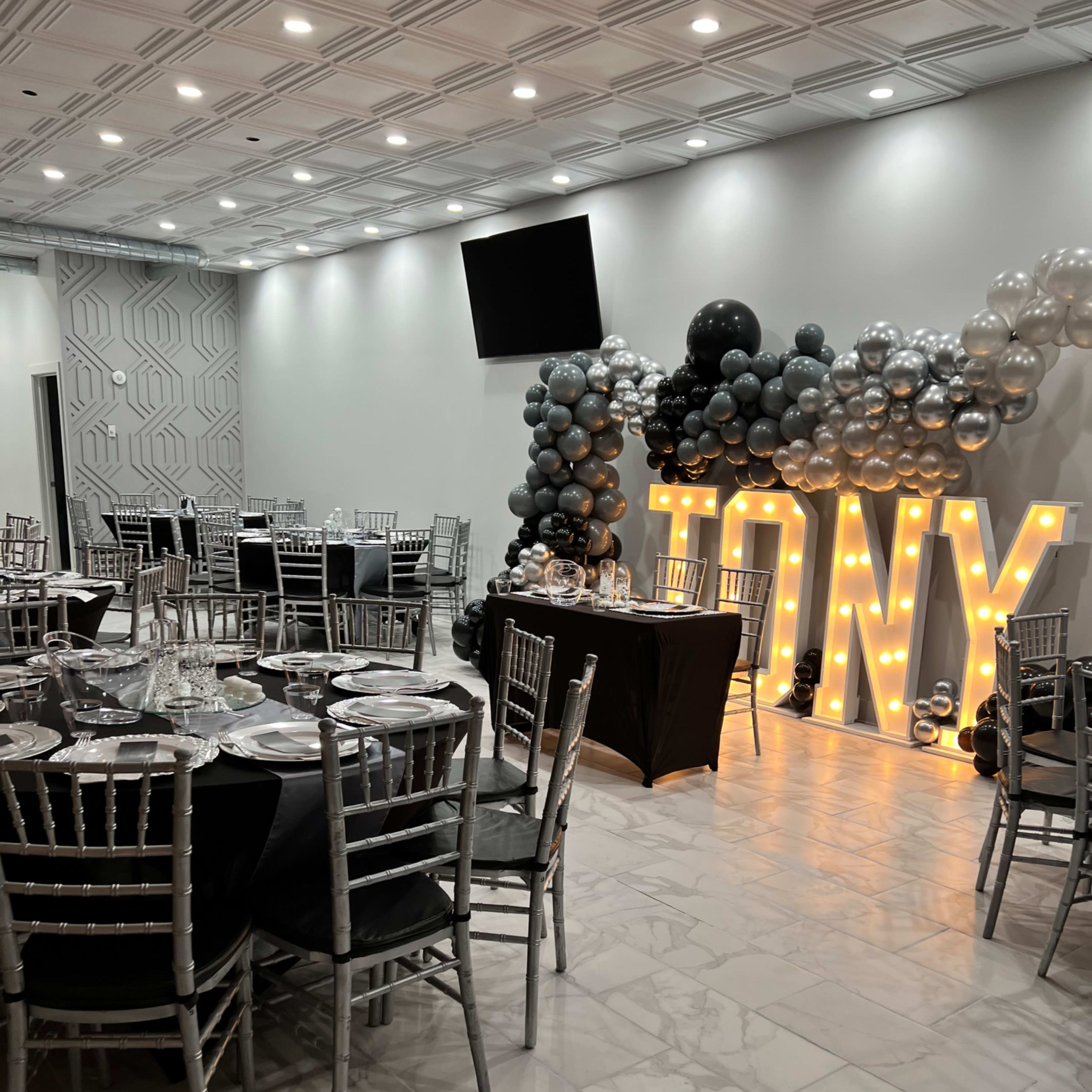 A decorated event space with tables set for a celebration, featuring silver and black balloons and illuminated letters spelling "TONY."