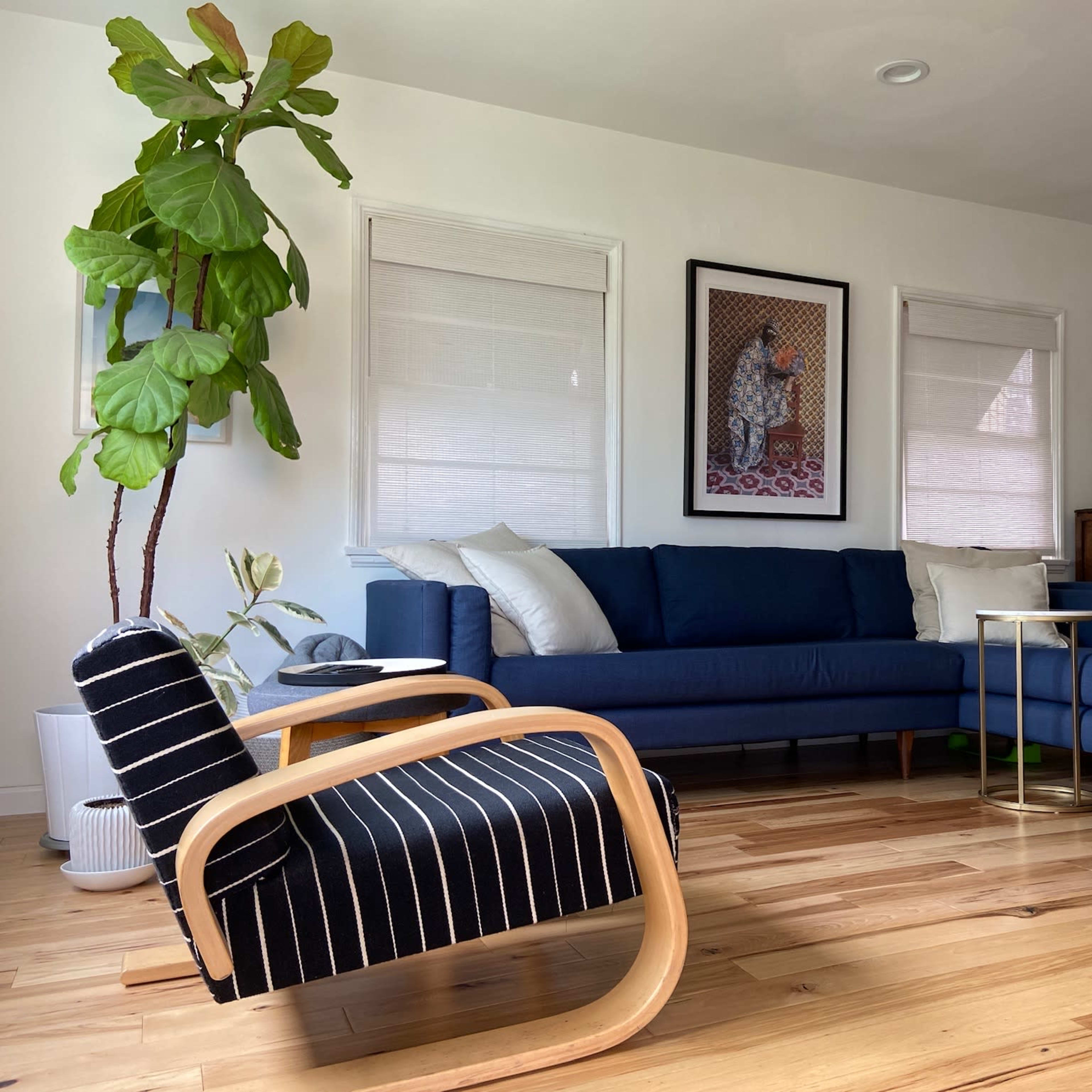 A blue sectional sofa and a striped armchair are positioned in a well-lit living room with hardwood floors and decorative artwork on the walls.