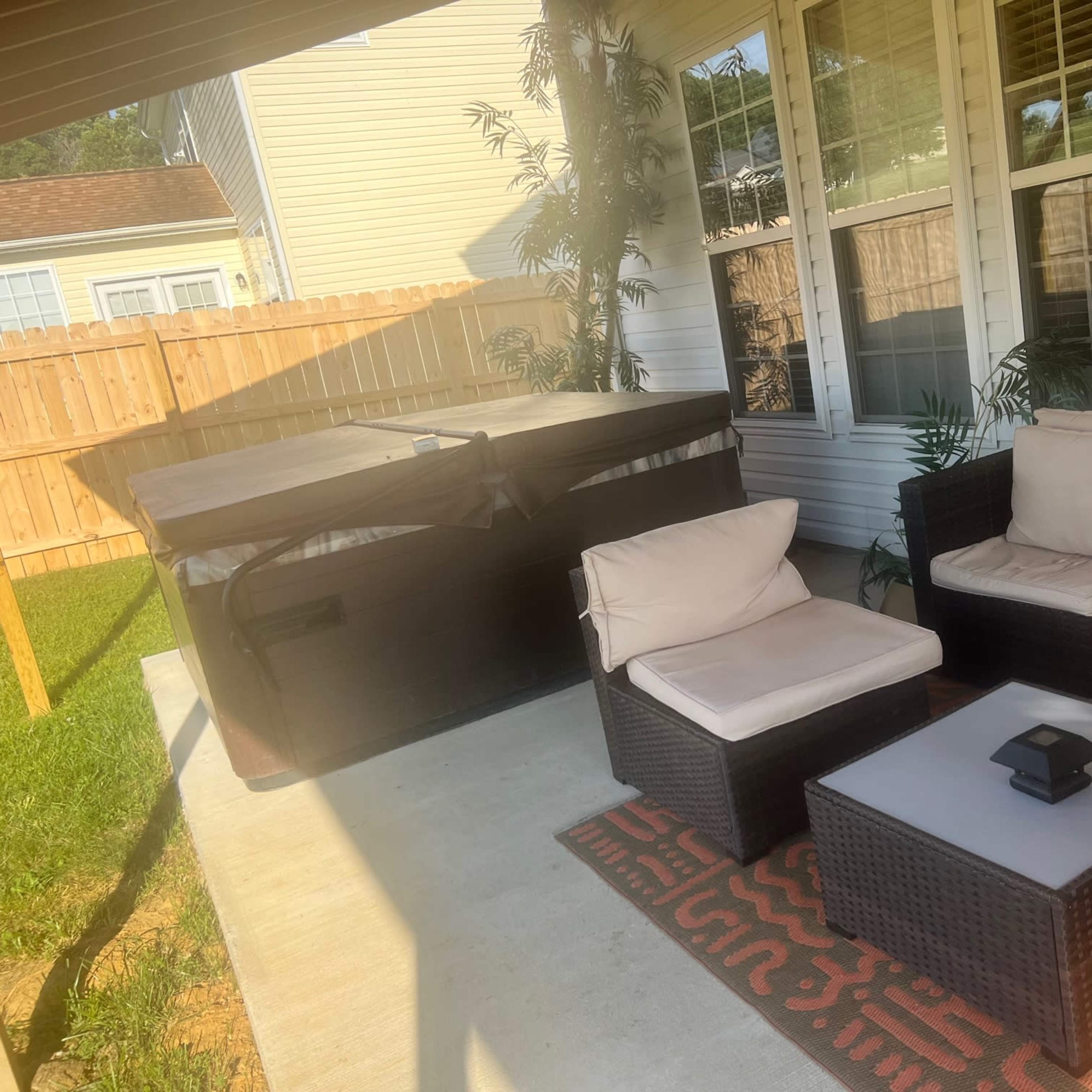 A patio with a beige sectional sofa, a small table, and a hot tub covered with a tarp, surrounded by a wooden fence and grass.