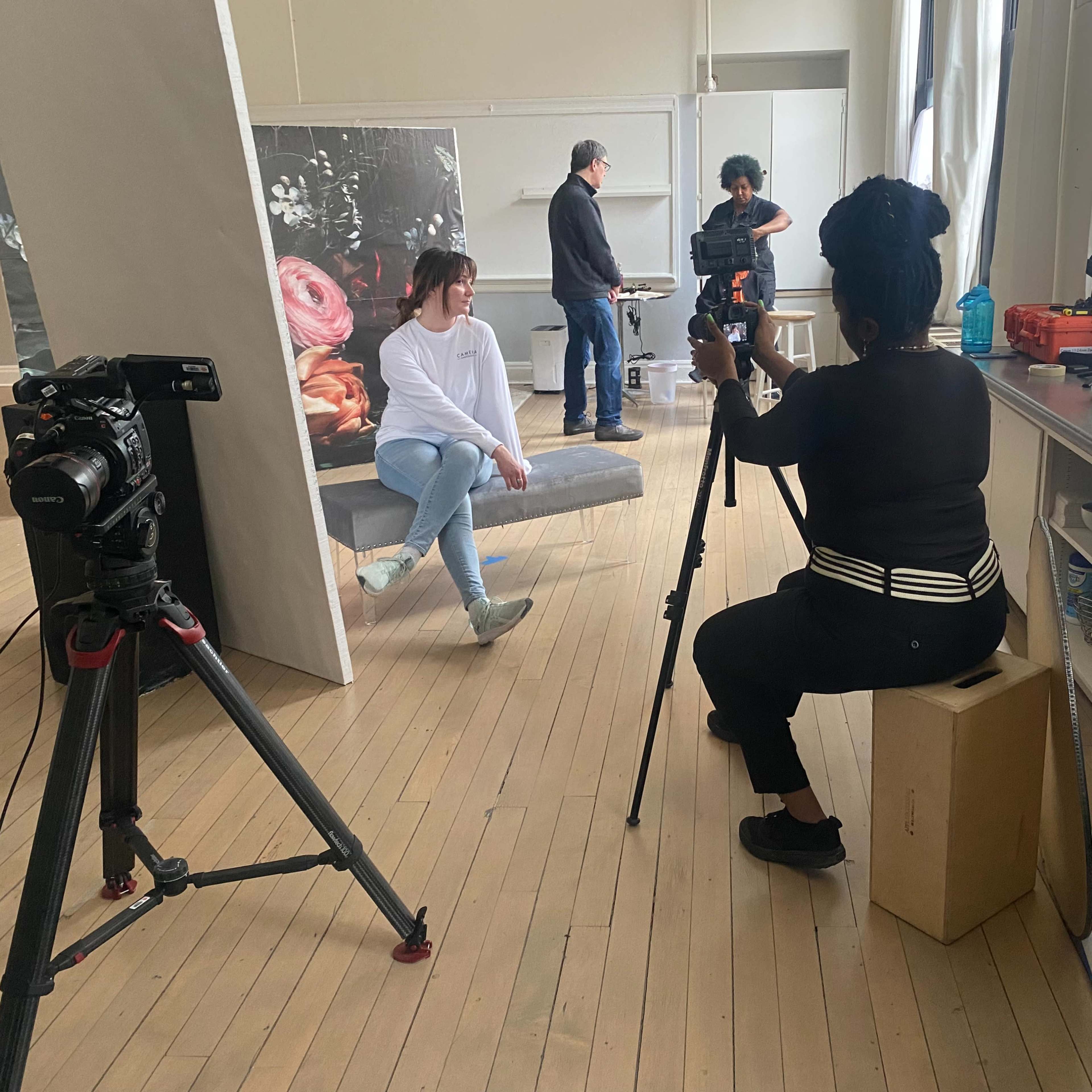 A woman sits in front of a camera while another person poses on a platform in a studio setting.