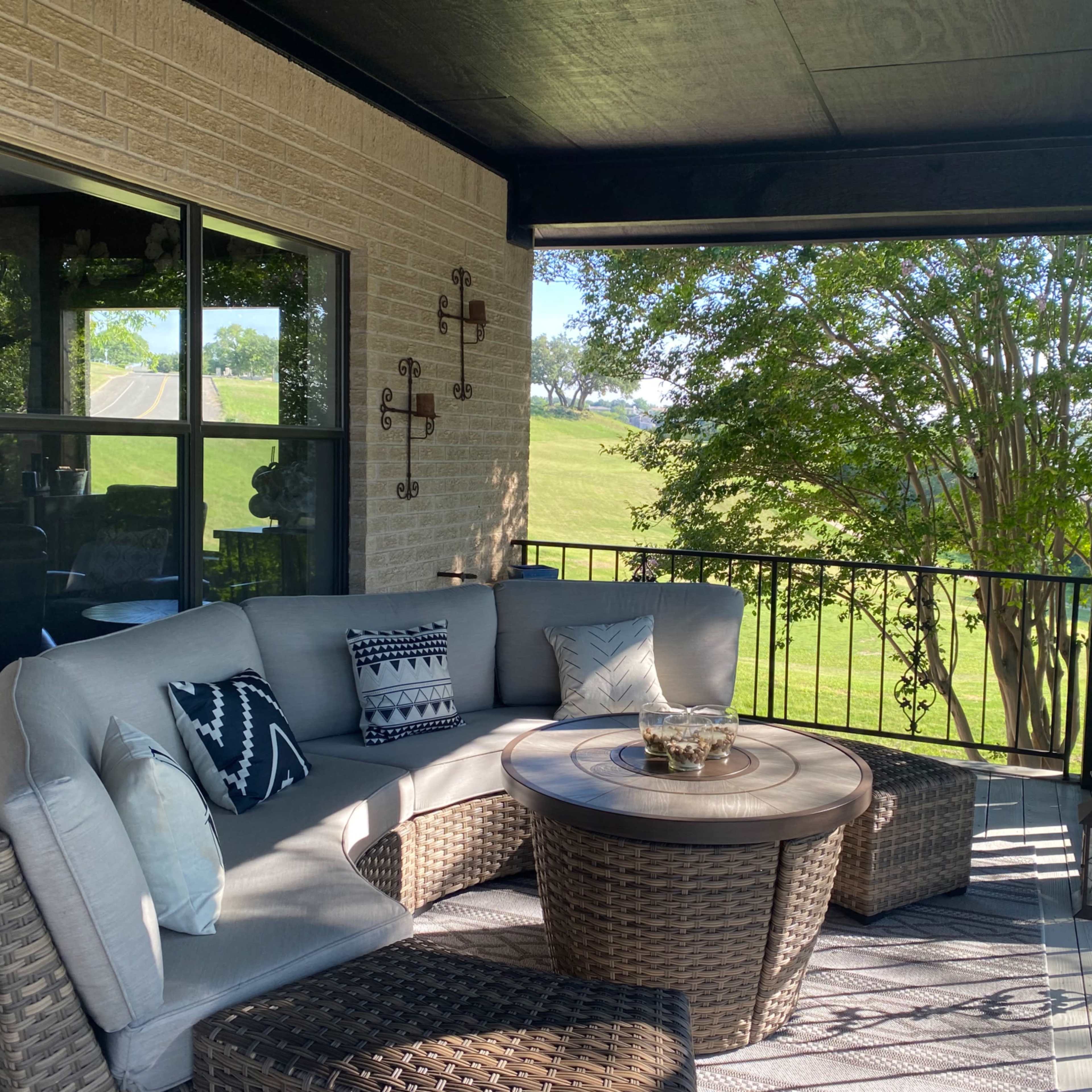A spacious outdoor balcony features a sectional couch and a round table, surrounded by greenery and a distant view of a hillside.