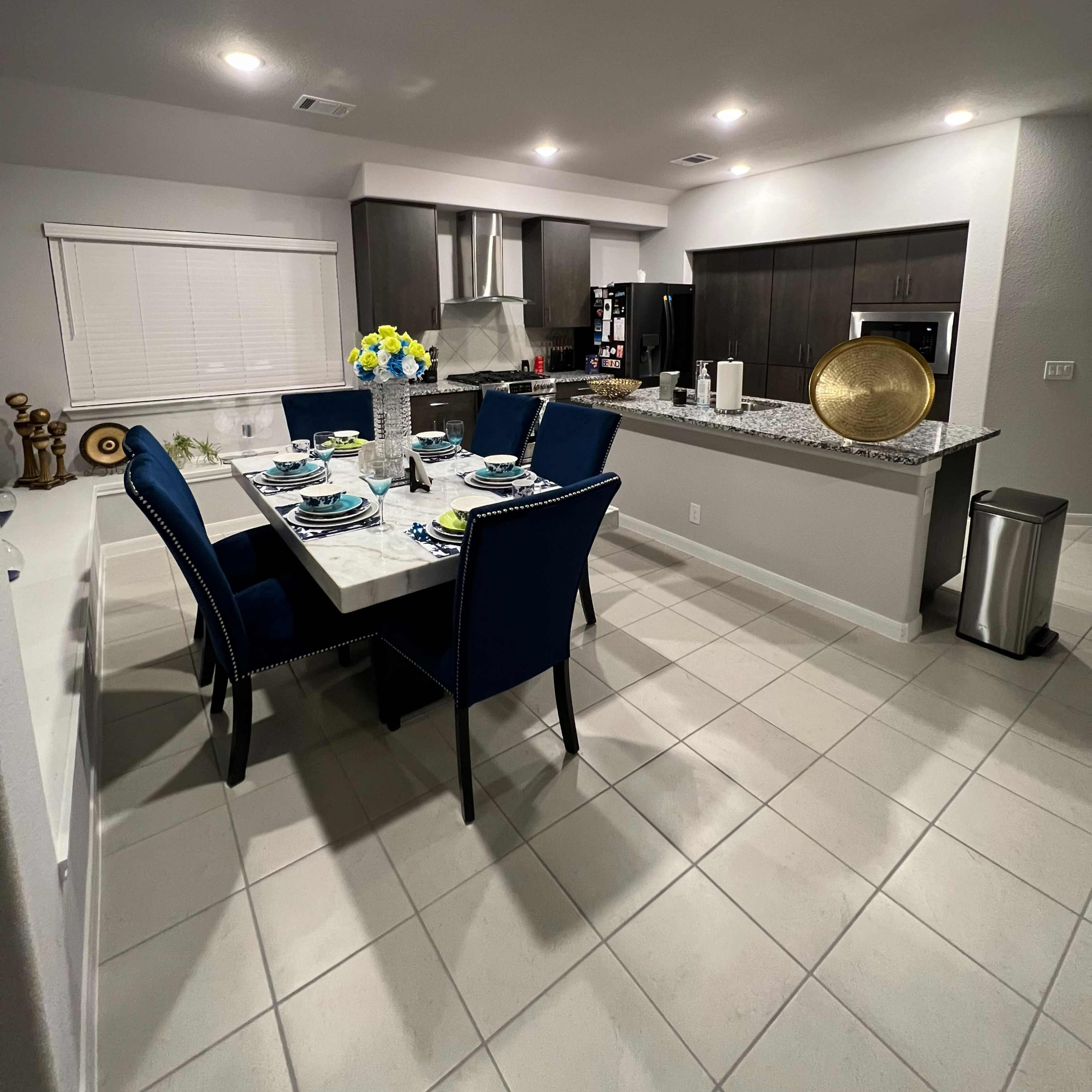 A modern kitchen and dining area with a table set for six, dark cabinets, and a countertop featuring decorative items.