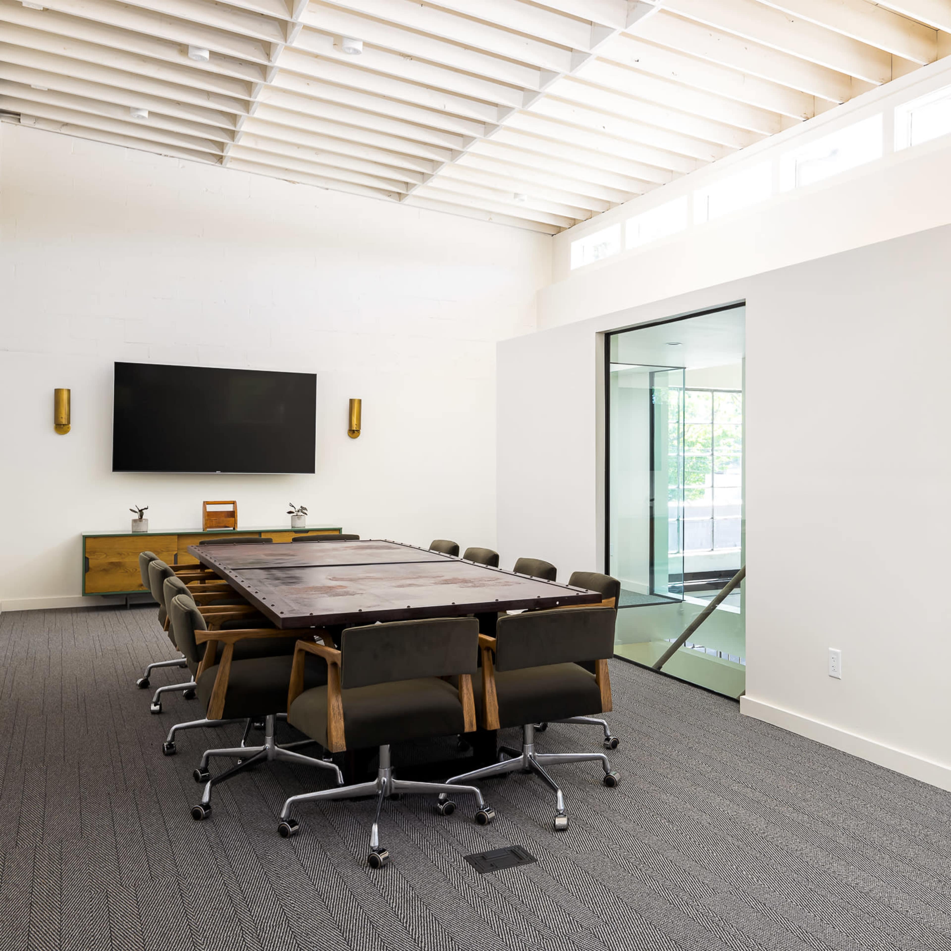 A modern conference room with a large table, ergonomic chairs, and glass-walled offices on either side.