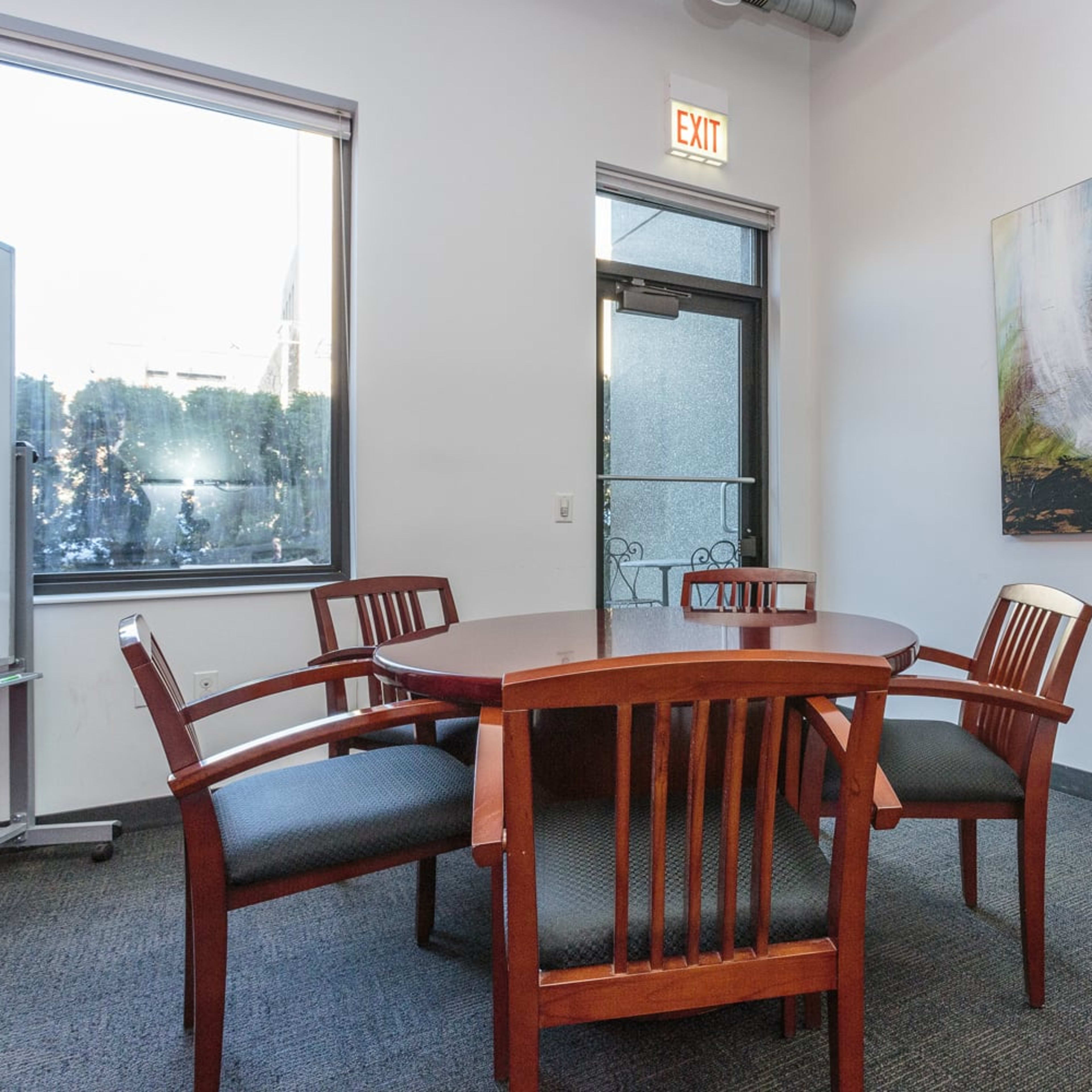 A meeting room features a round wooden table surrounded by four chairs, a whiteboard on wheels, and large windows letting in natural light.