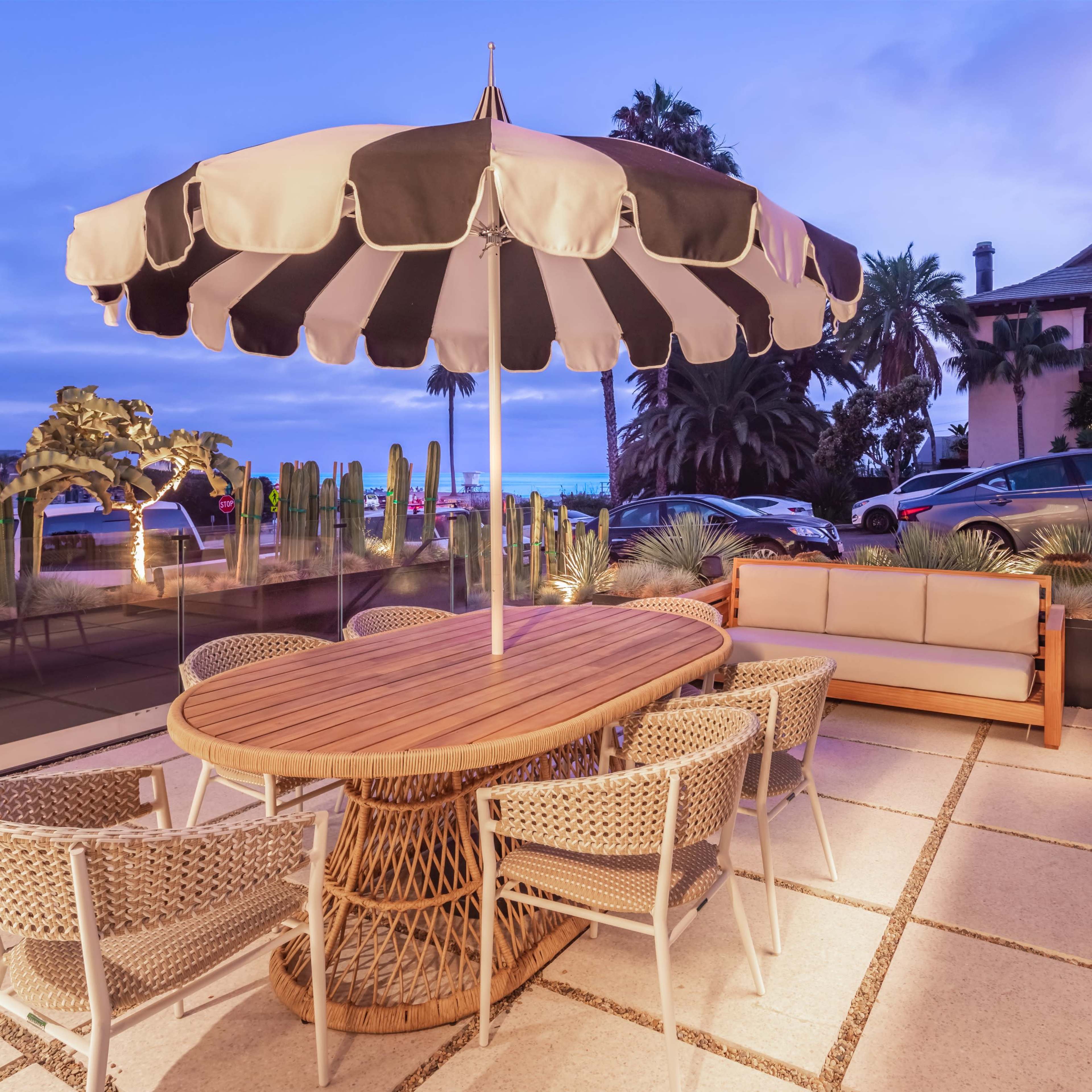 A round wooden table with woven chairs is set under a striped umbrella on a patio surrounded by palm trees and a view of cars parked nearby.