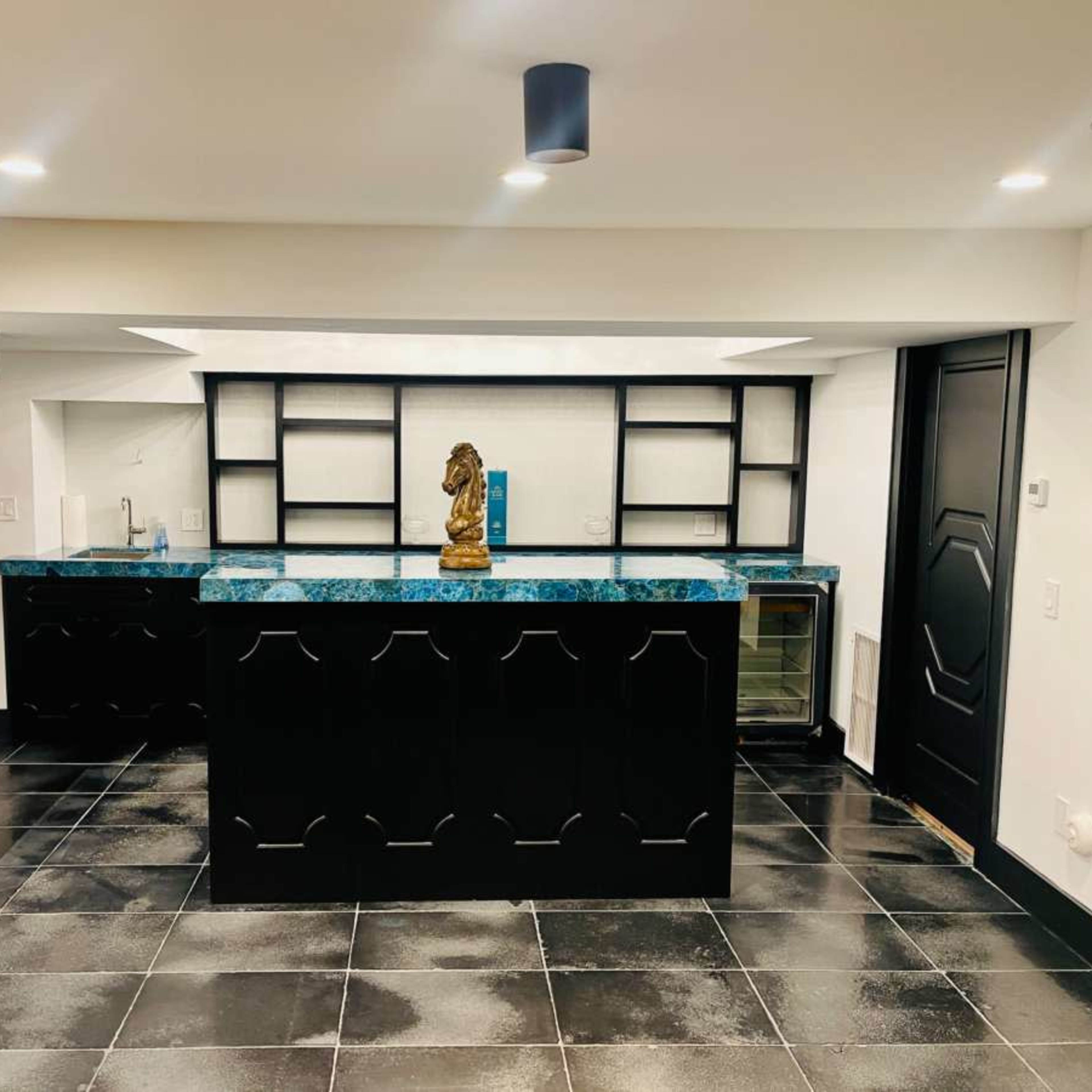 A modern basement bar area with a blue granite countertop, black cabinetry, and a decorative statue on the counter.