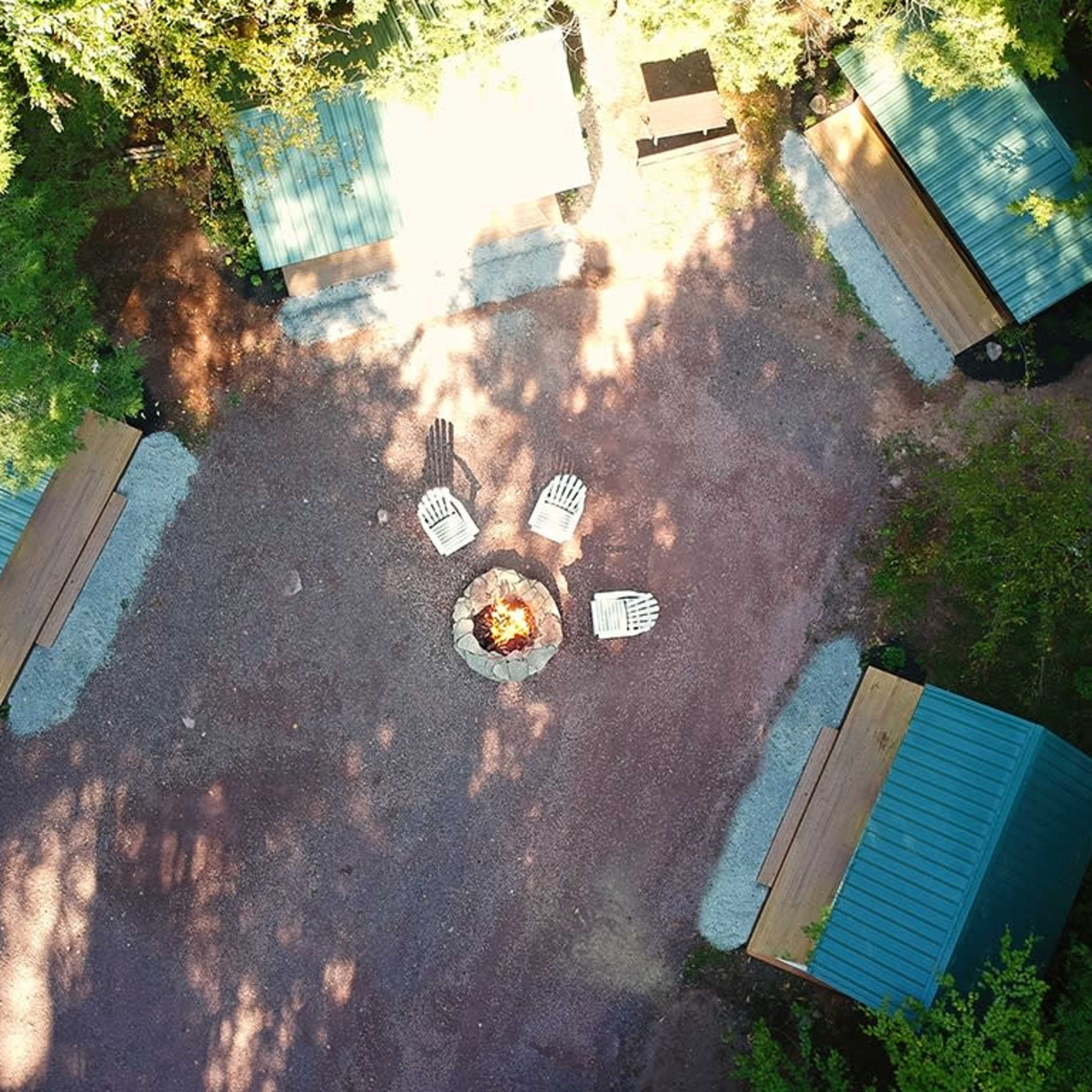 An aerial view of a circular campsite with several metal-roofed cabins surrounding a fire pit and arranged seating.