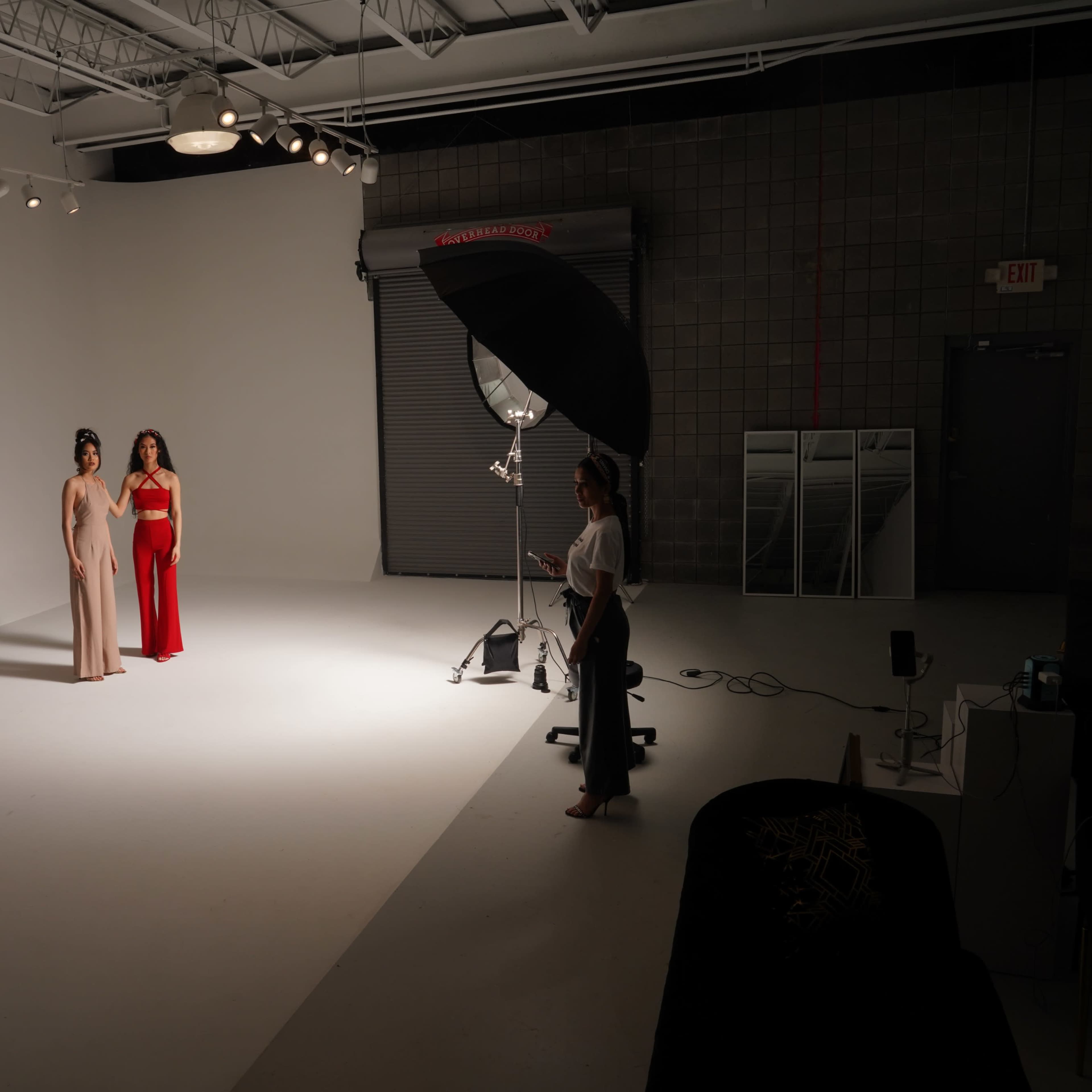 Two models stand in a well-lit studio space, with one wearing a beige outfit and the other in red, while a photographer positions a large softbox light.
