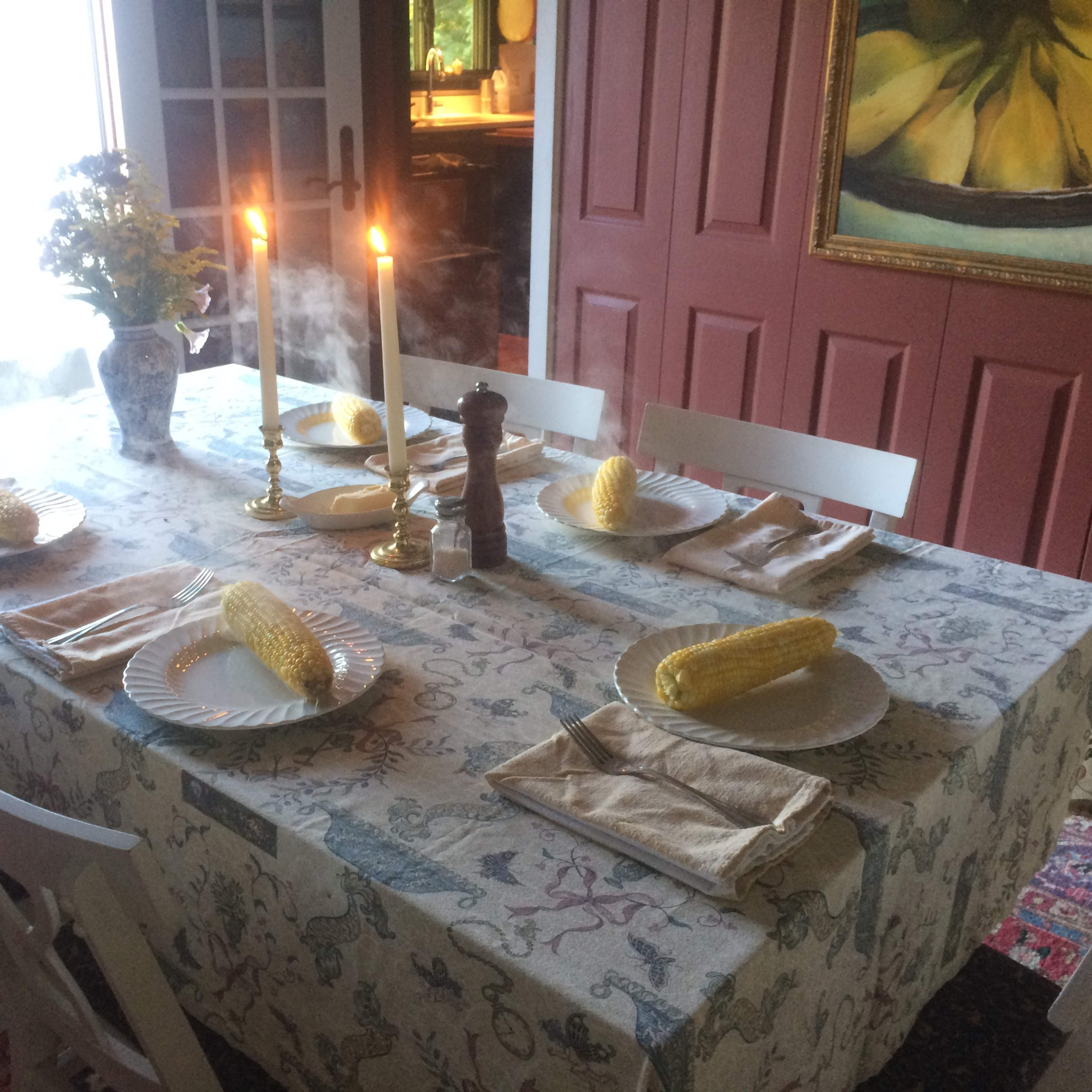 A dining table is set with plates of corn, utensils, and lit candles, surrounded by chairs and decorated with a floral vase.