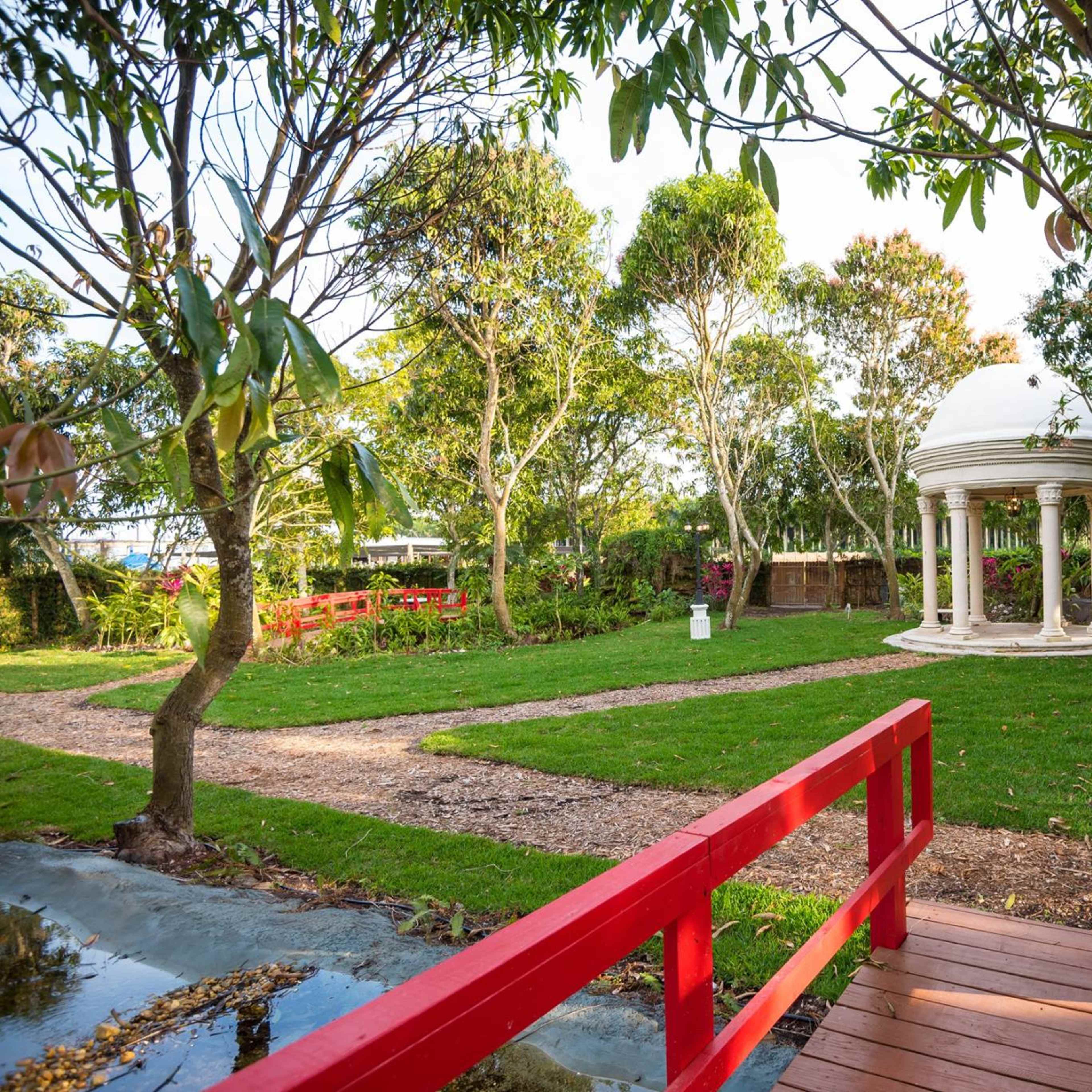 A red wooden bridge leads to a white gazebo surrounded by lush greenery and trees in a landscaped garden.