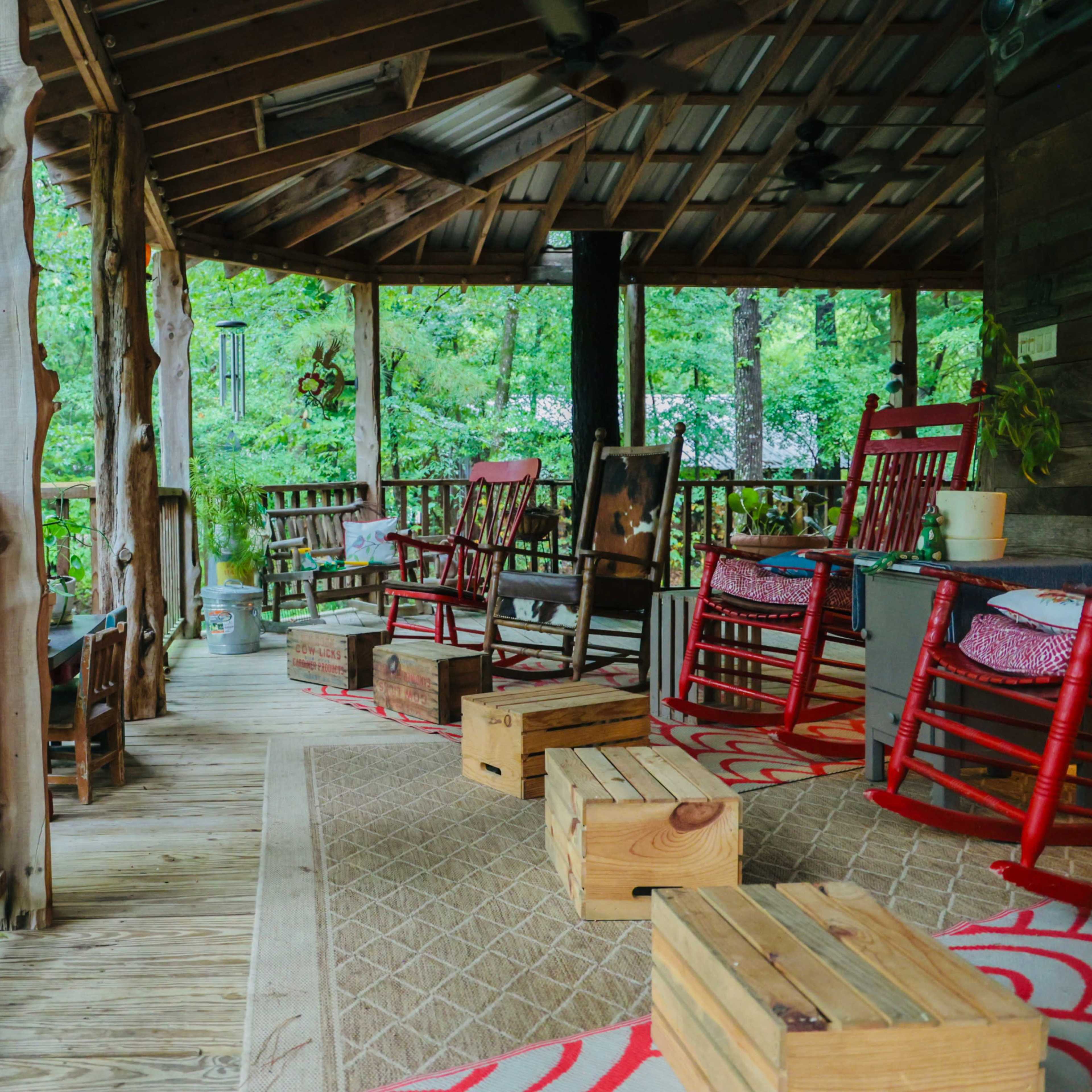 A wooden porch with red rocking chairs, a table, and wooden stools, surrounded by greenery.