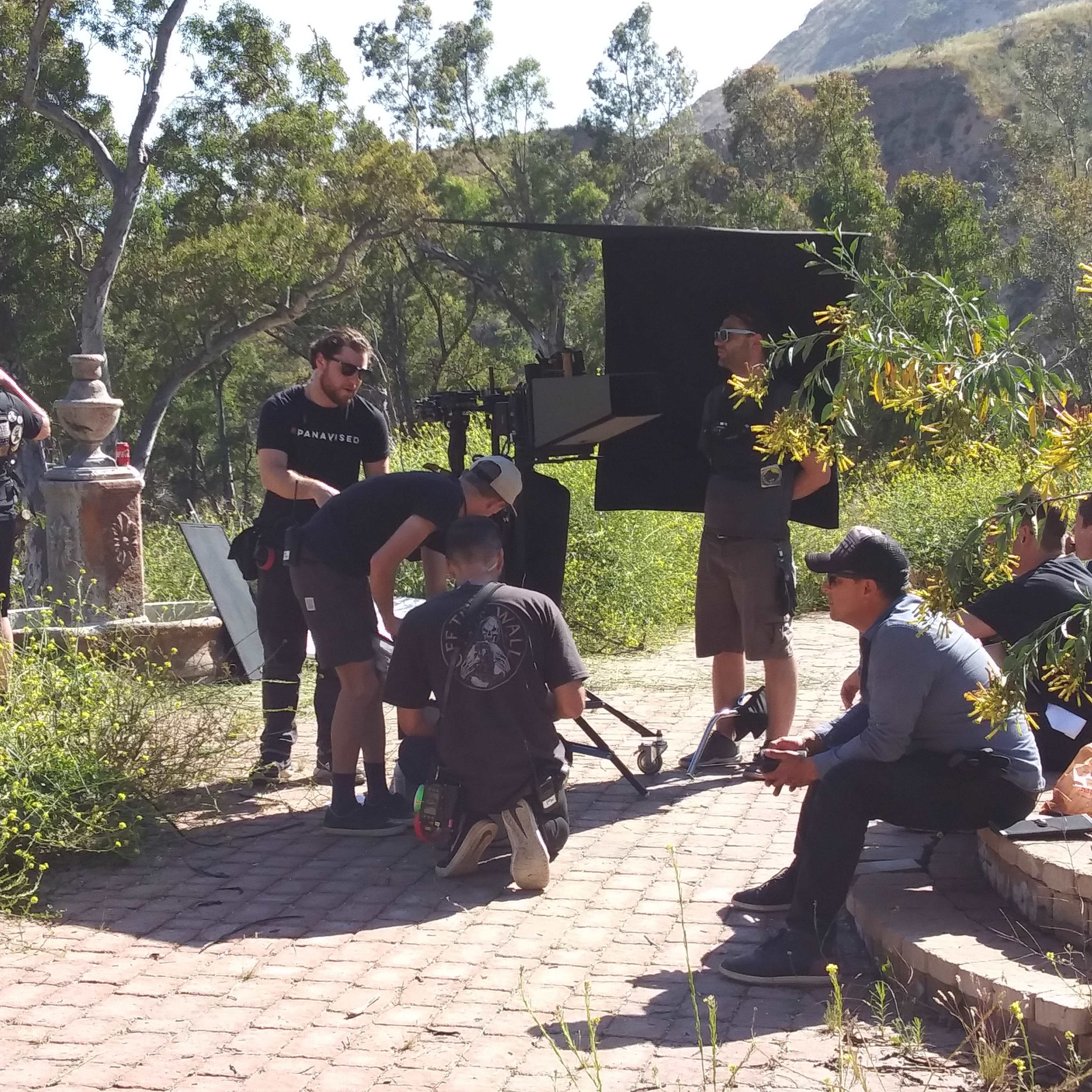 A film crew sets up equipment in a natural outdoor location surrounded by trees and greenery.