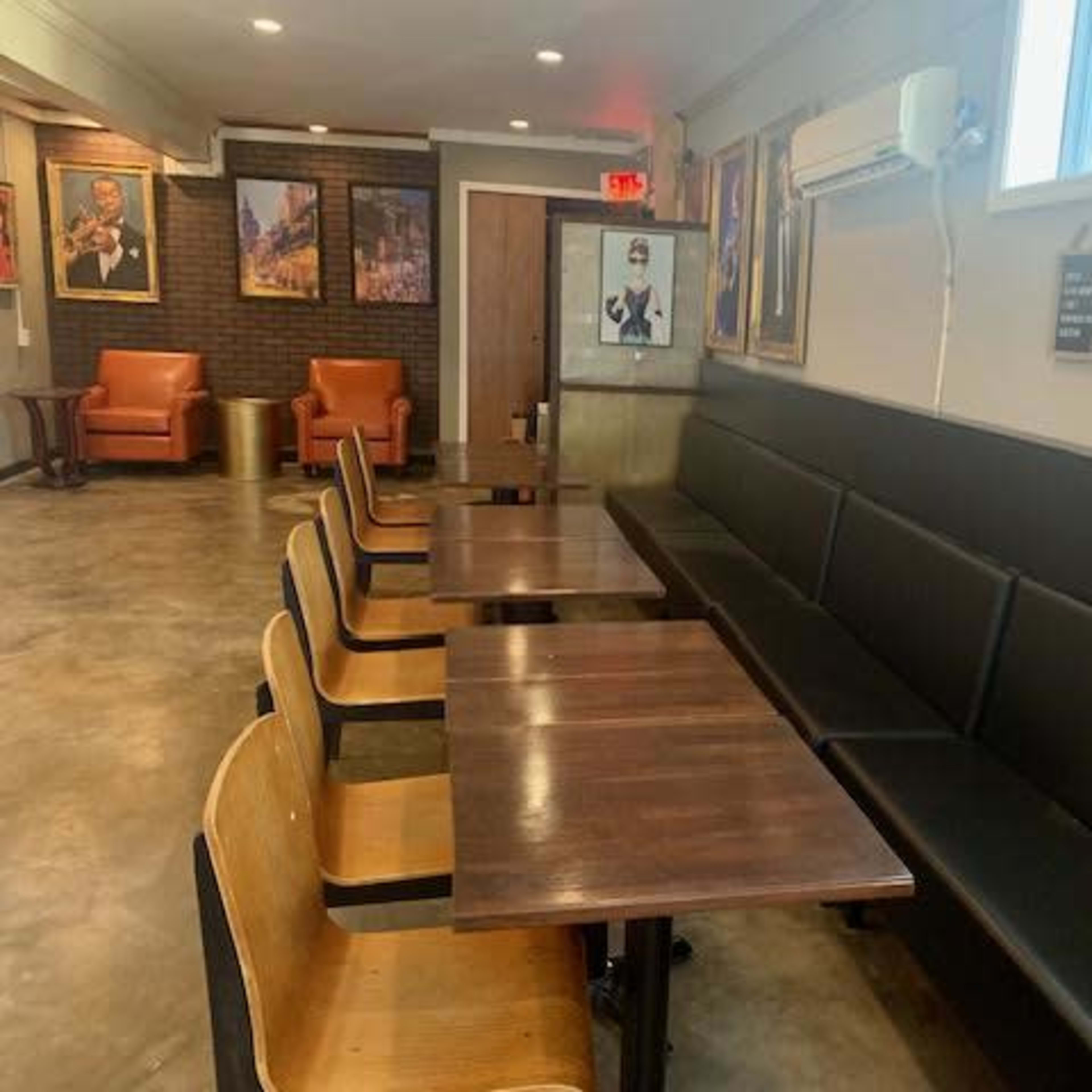 A café interior featuring wooden tables and black cushioned seating against a wall with framed portraits and a window.