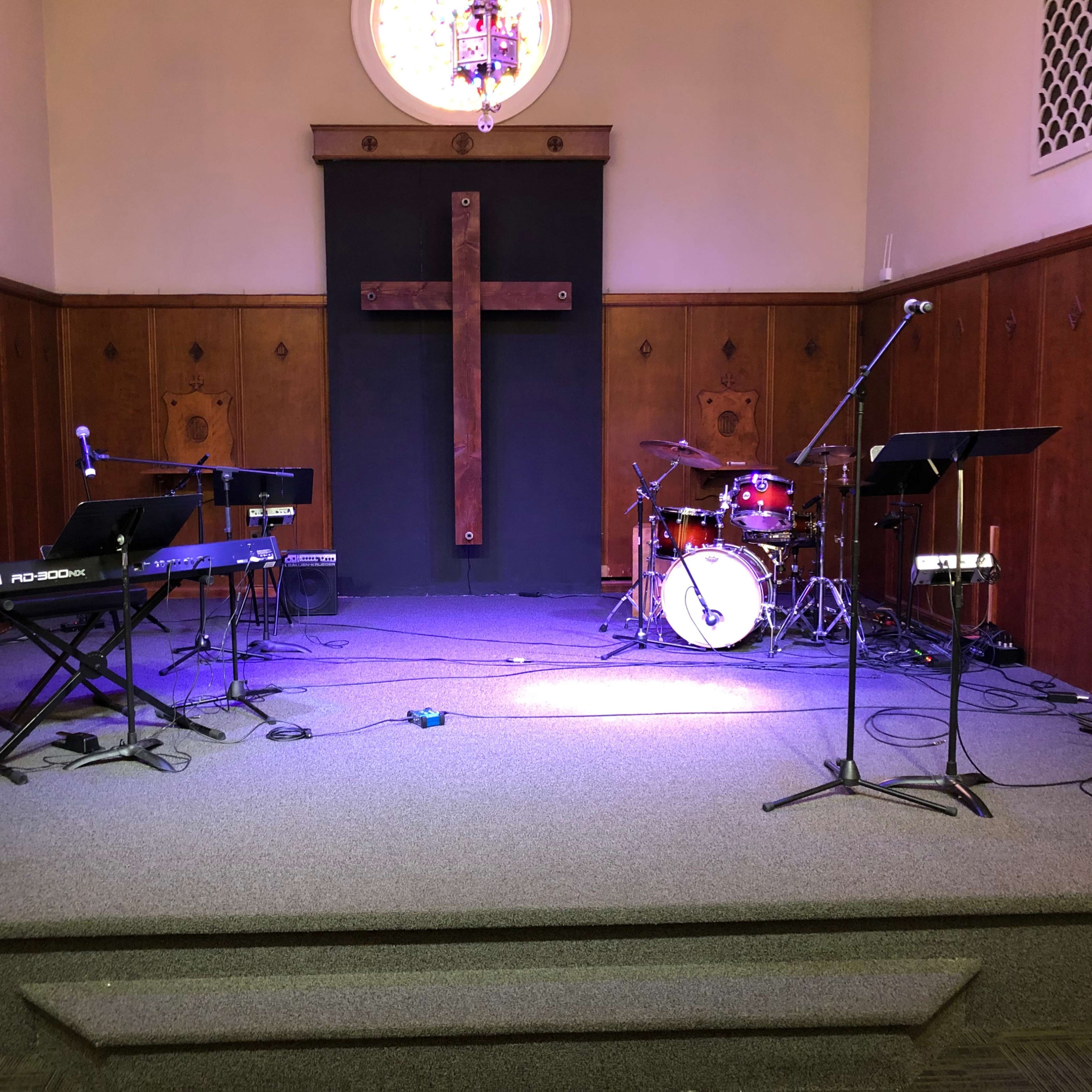 A stage setup in a church featuring musical instruments, including a keyboard, microphones, and a drum set, with a large wooden cross on the wall behind.