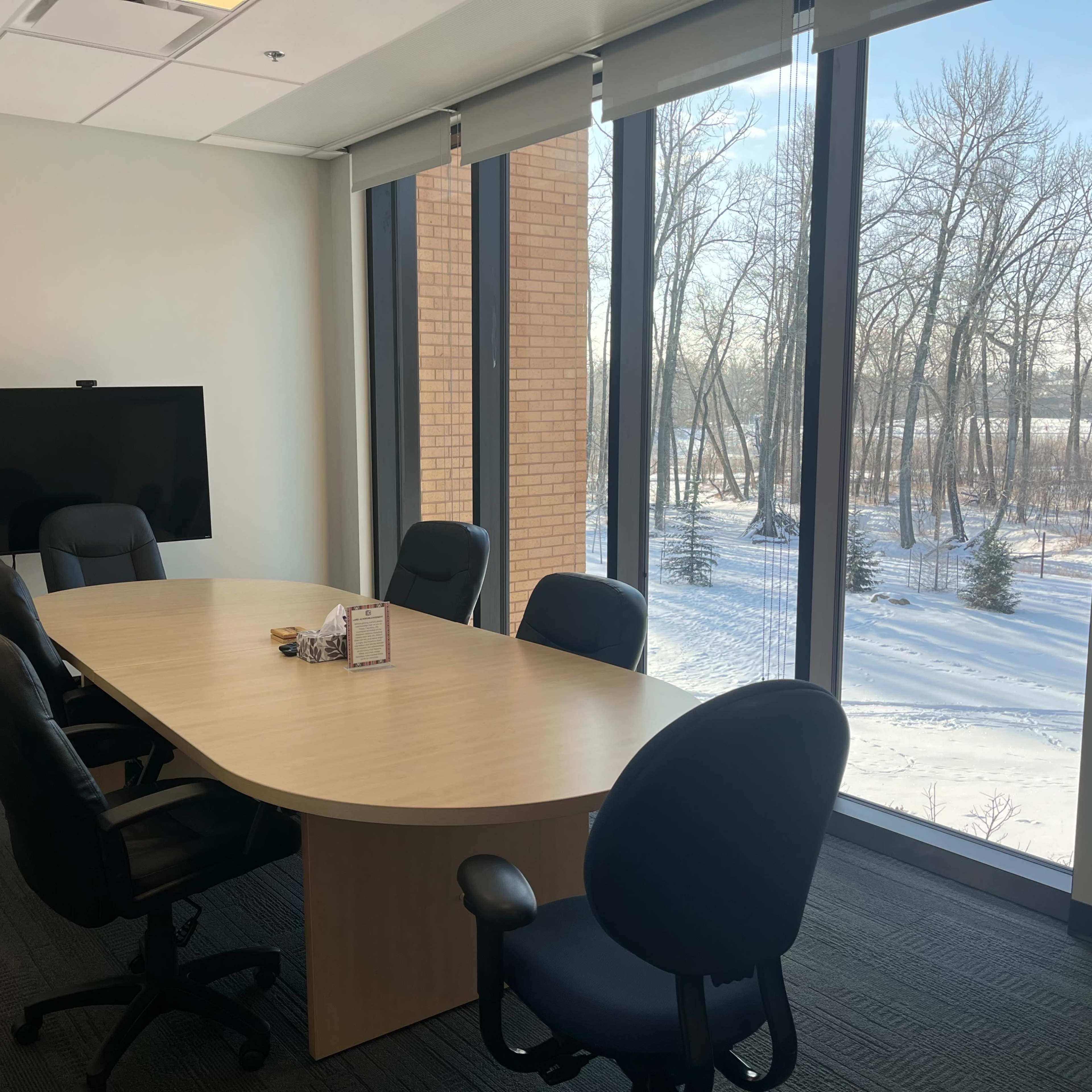 A modern conference room with a large wooden table surrounded by six black office chairs and a large window overlooking a snowy landscape with trees.